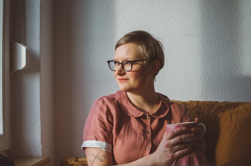 Marcia sitz auf einer Couch, mit einer Tasse in der Hand und schaut aus einem Fenster