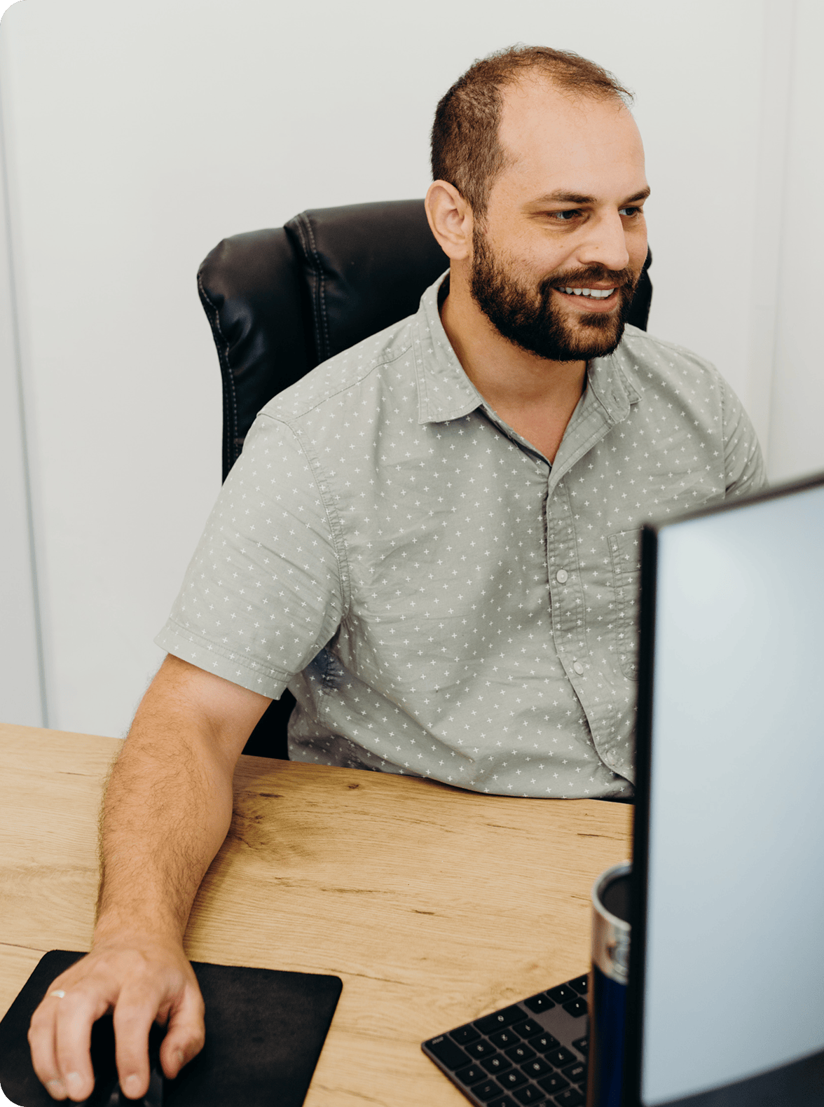 An adult man with a short beard sits at a wooden desk in an office, smiling while looking at a computer monitor. He wears a light gray short-sleeve shirt with a subtle dot pattern; a keyboard and mouse rest on the desk beside the monitor.