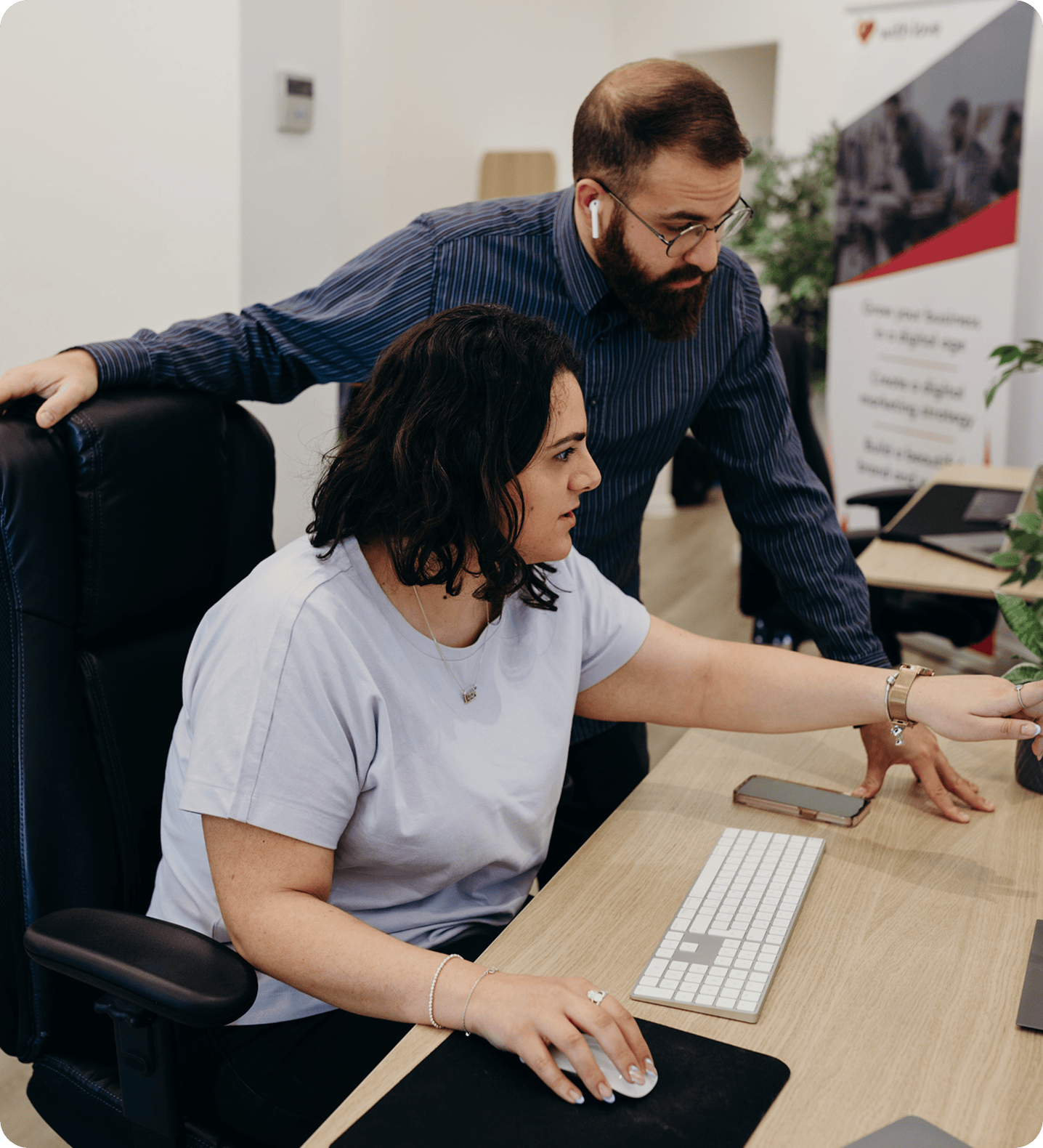 Bearded man leaning over a desk guides a seated woman as she works on a computer. They’re in a modern office with a keyboard, monitor, and a plant visible in the background.