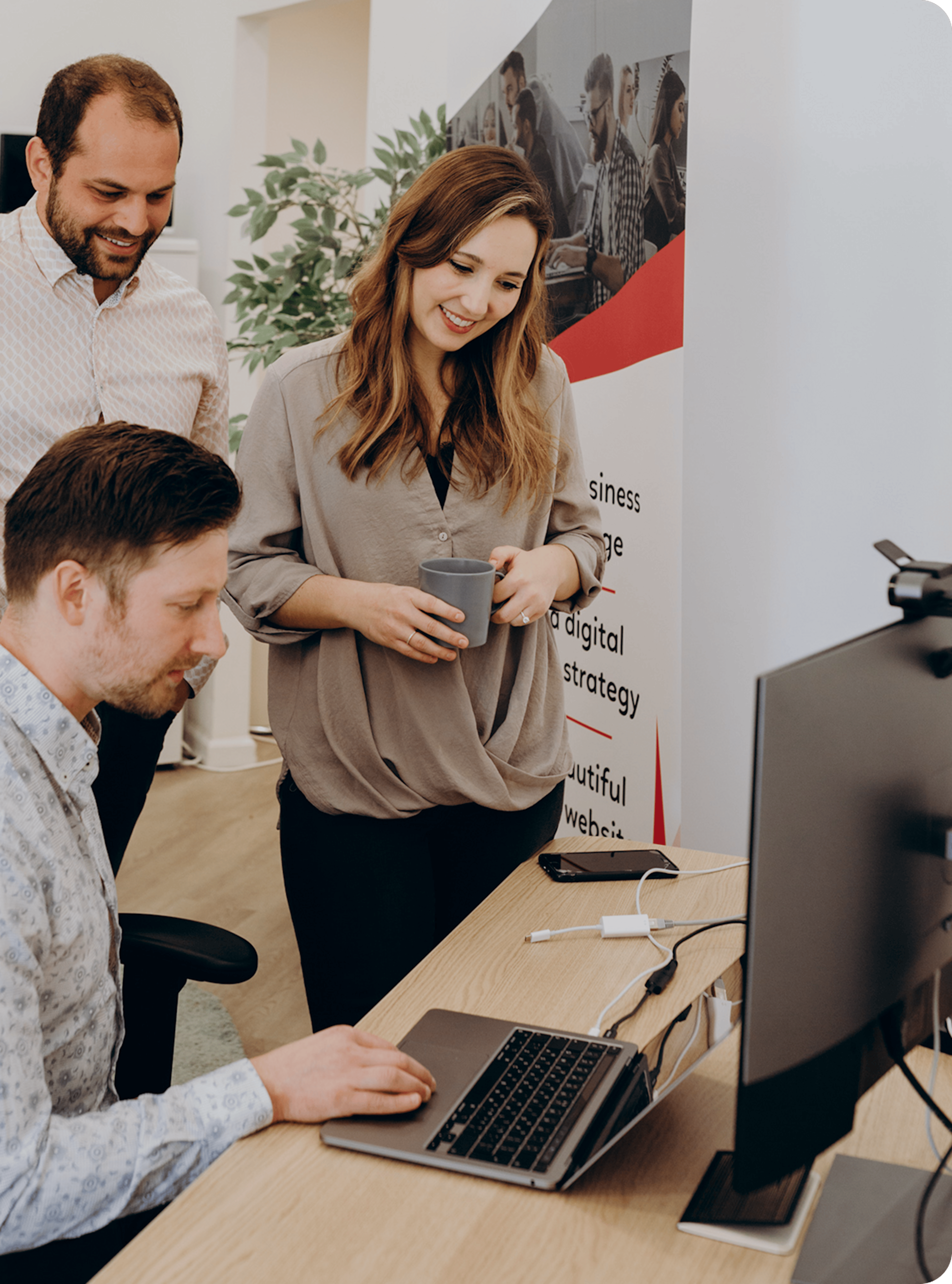 Three coworkers collaborate at a desk with a laptop and a large monitor; the woman in the center holds a coffee cup and smiles as the two men look on, in a bright office with a banner and a plant in the background.