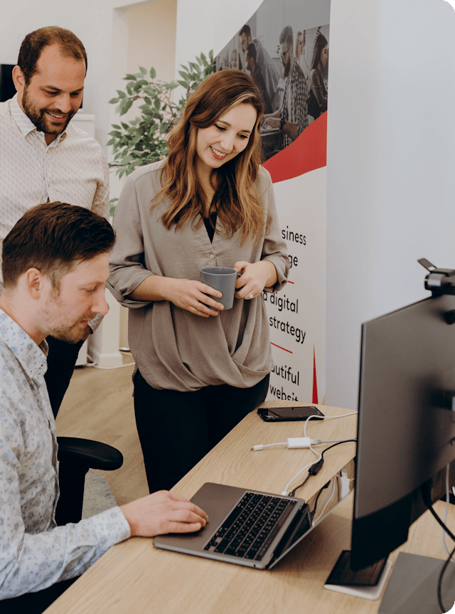 Three coworkers collaborate at a desk with a laptop and a large monitor; the woman in the center holds a coffee cup and smiles as the two men look on, in a bright office with a banner and a plant in the background.