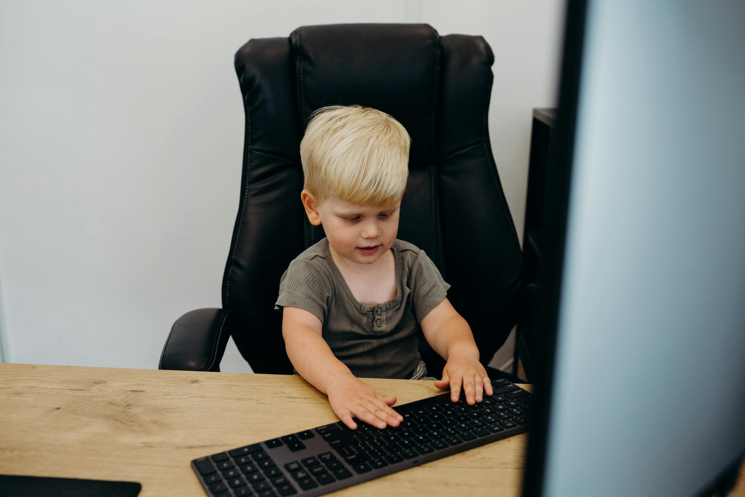 A young blonde toddler sits in a tall black leather office chair at a wooden desk, focused on typing on a full-sized keyboard in front of a computer monitor, suggesting a playful moment in a digital marketing workspace.