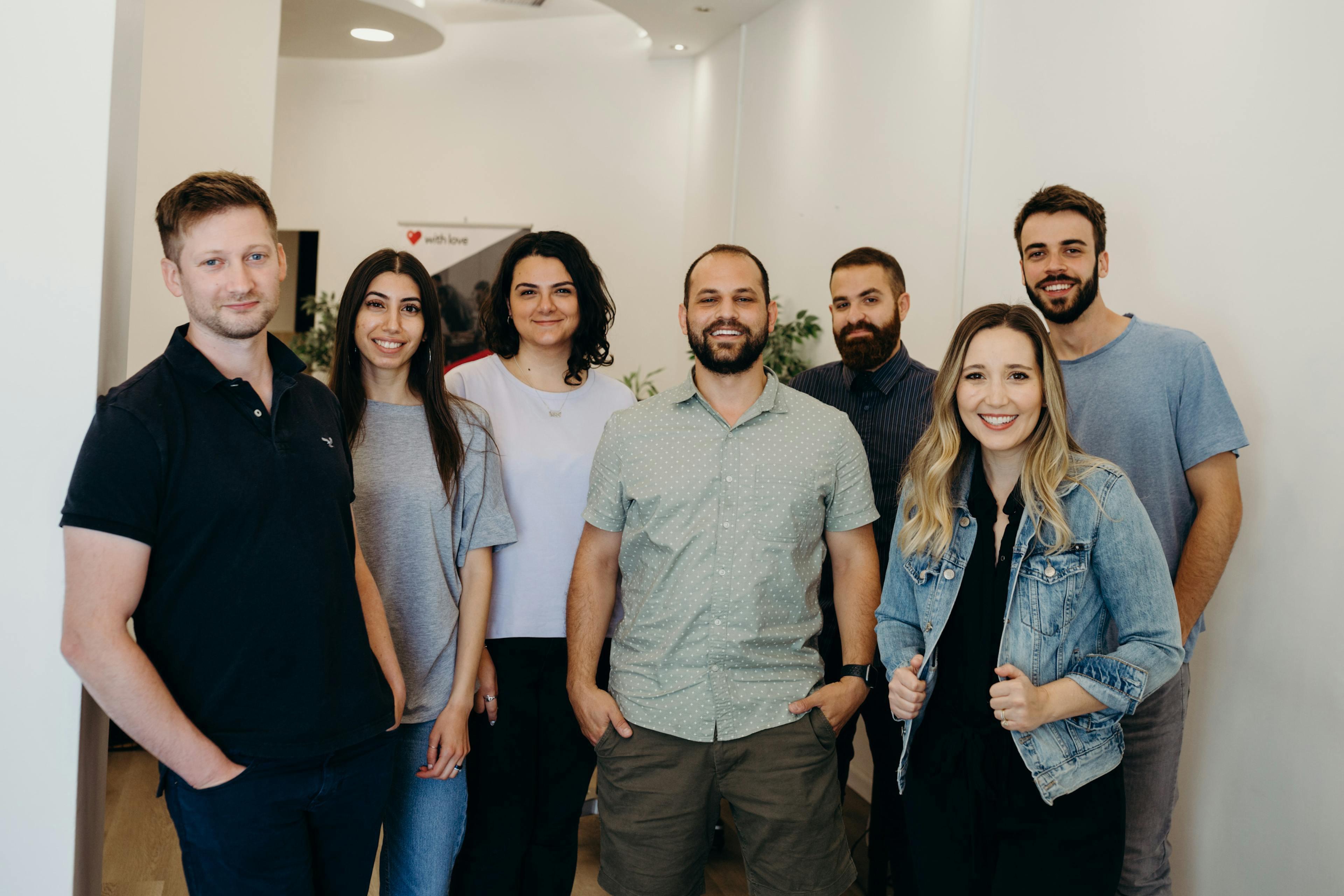 Seven diverse people stand together in a bright office hallway, smiling at the camera. They wear casual clothing, with a man in a light green shirt centered among six others.