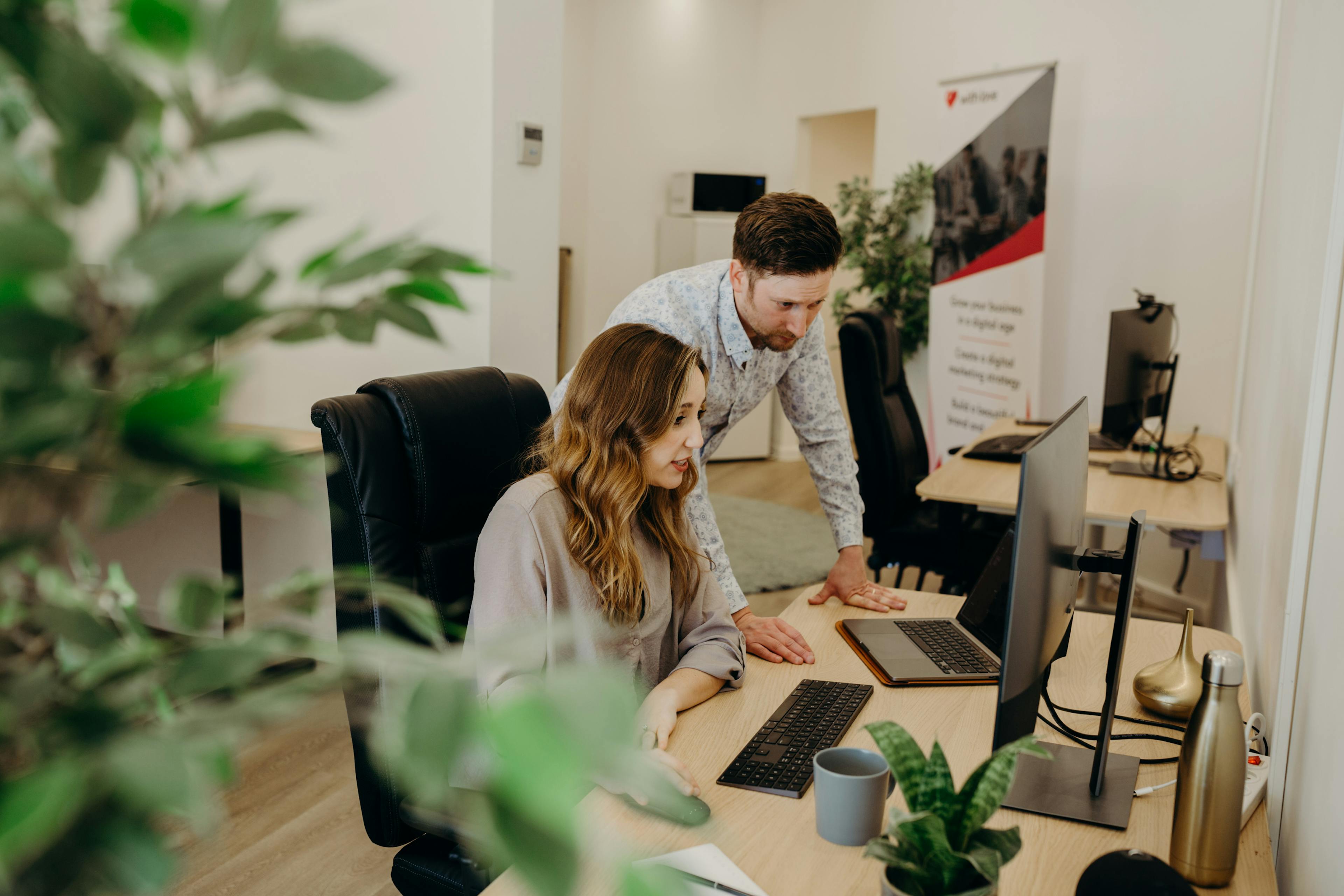 Two coworkers work at a bright office desk: a woman seated at a laptop while a man leans over to assist, with an additional monitor, mug, and small plants nearby, and a banner in the background.