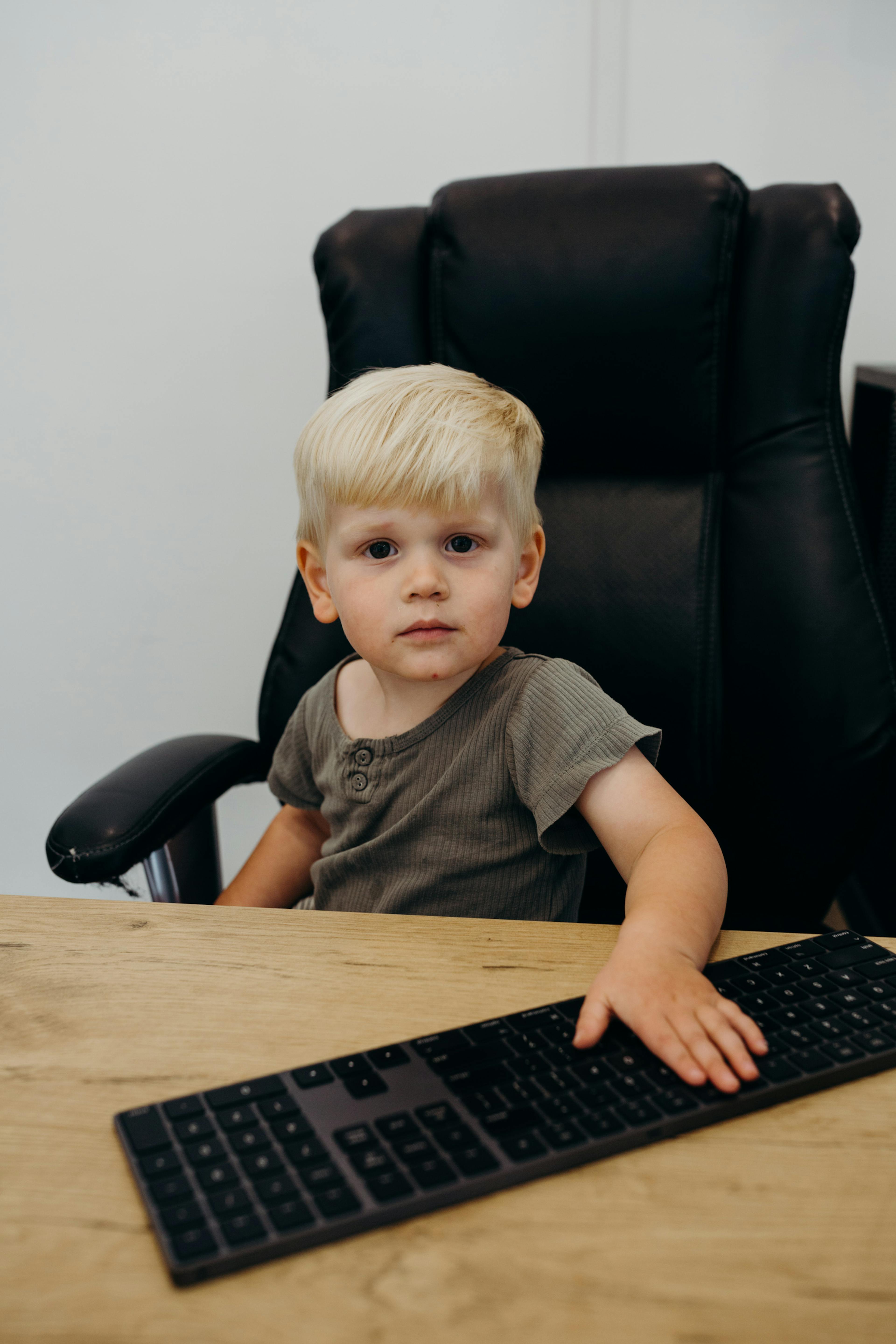 A young blond-haired child sits in a black office chair at a wooden desk, with one hand resting on a computer keyboard. He wears a brown shirt and looks toward the camera as the light gray wall frames the scene.