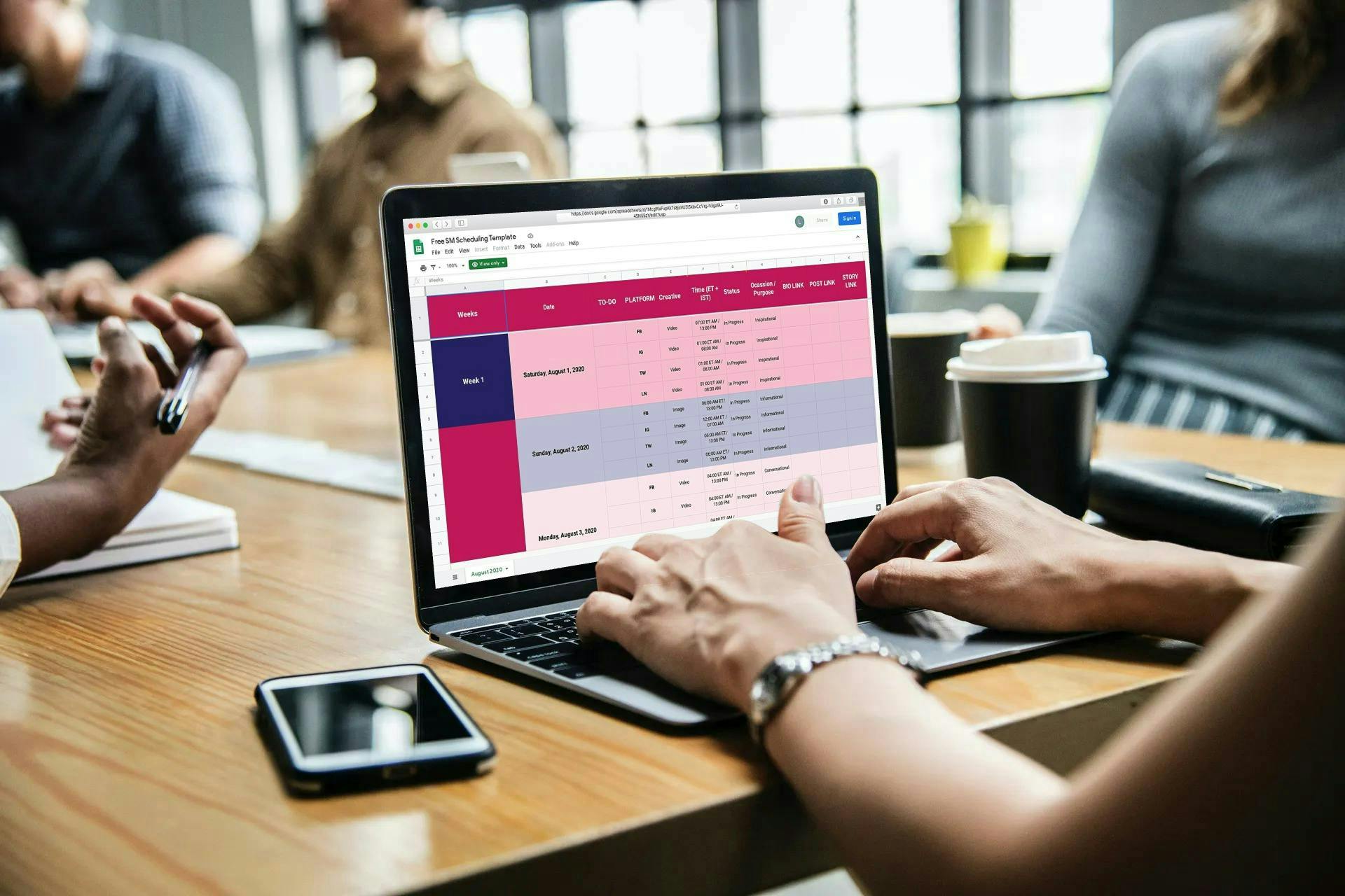 Hands typing on a laptop displaying a pink and purple data dashboard, while colleagues sit around a wooden table in a bright, modern office; a smartphone and a takeaway coffee cup are nearby.