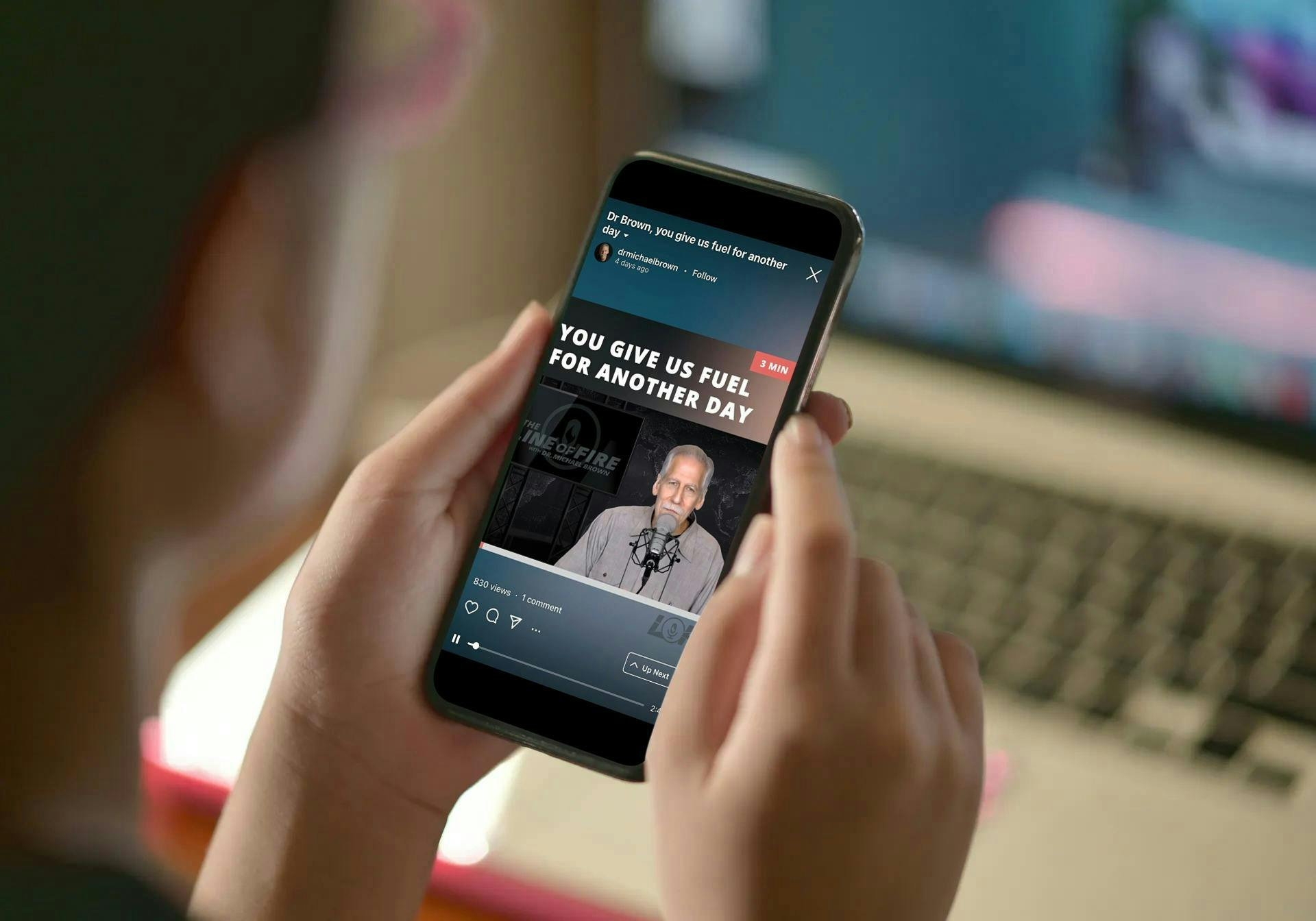 A person holds a smartphone displaying a video with the bold headline 'YOU GIVE US FUEL FOR ANOTHER DAY', showing a man speaking into a microphone in a studio; a blurred laptop keyboard is visible in the background.