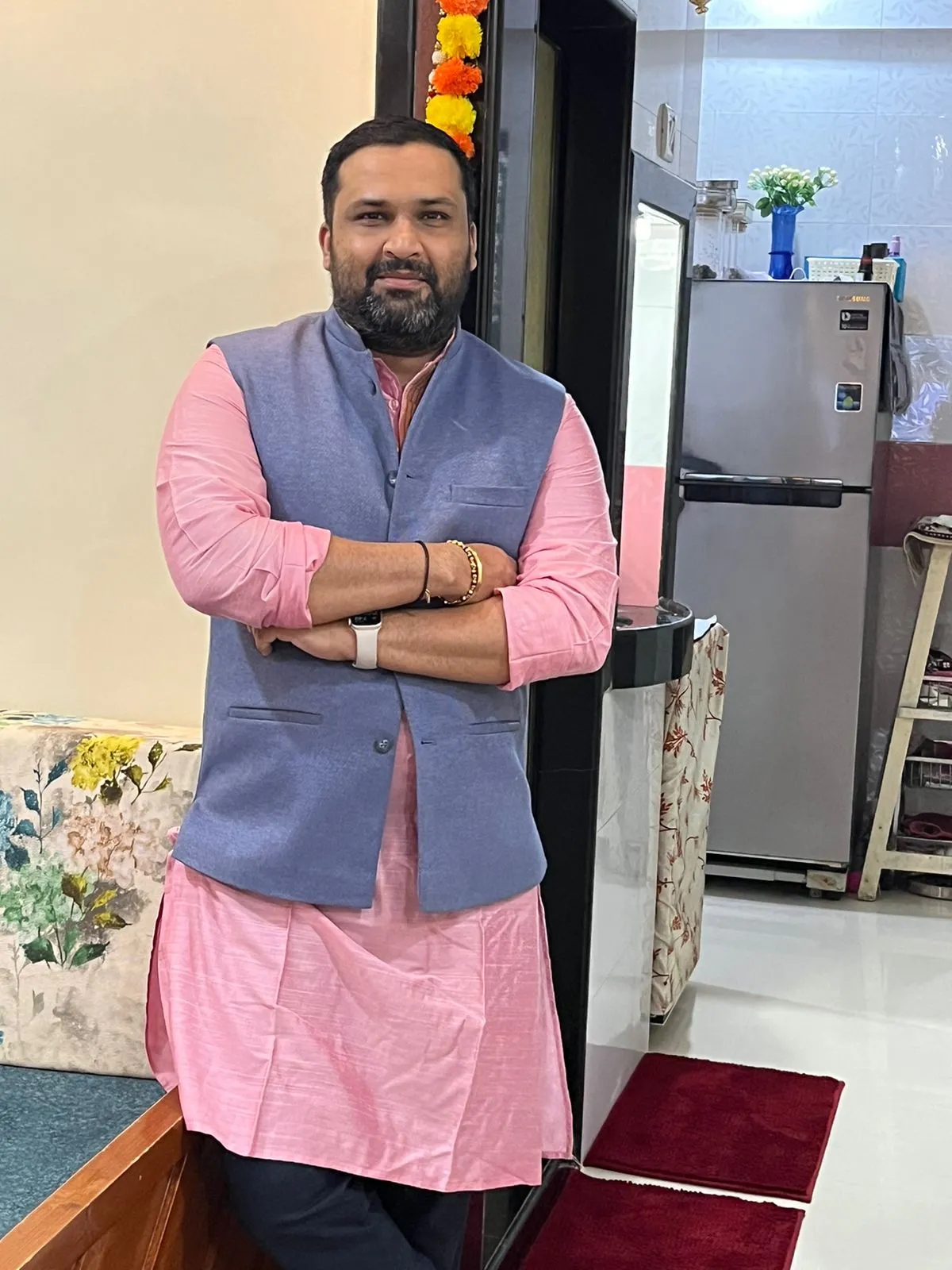 Bearded man wearing a pink kurta and a blue sleeveless vest stands with arms crossed in a home interior, beside floral upholstery and a refrigerator, with a festive garland visible at the doorway.