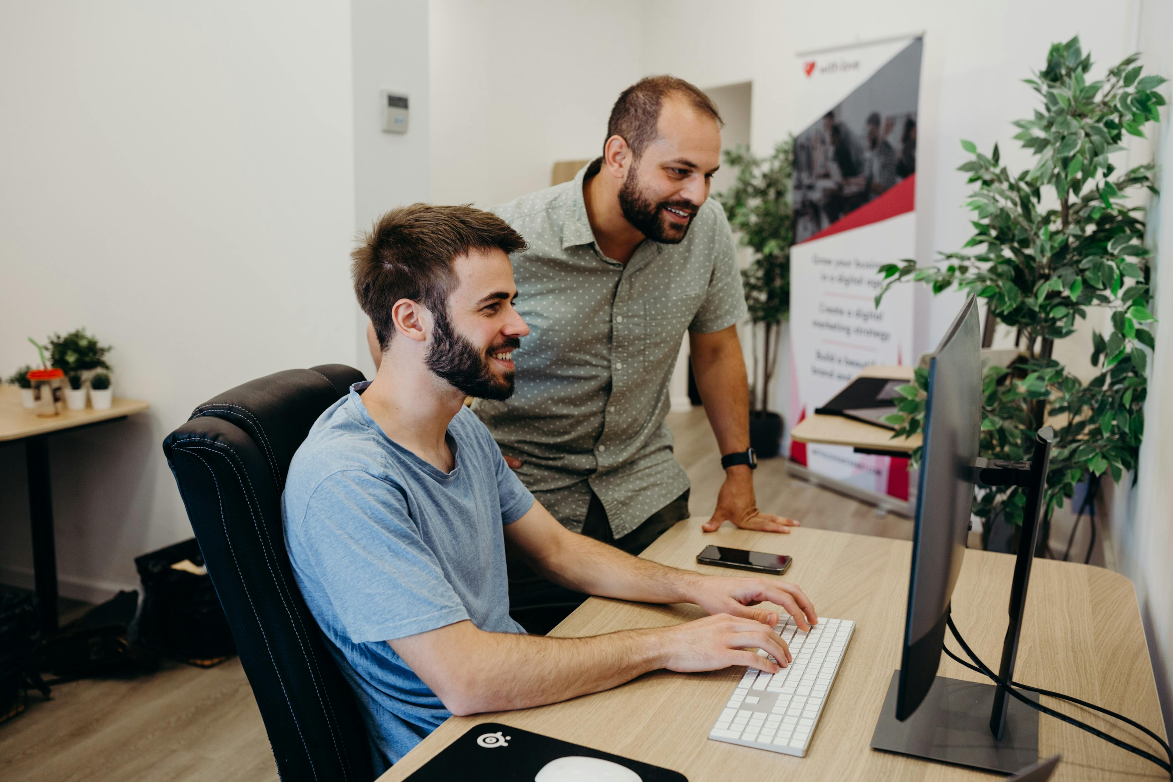 Two men in a bright office collaborate at a desk: one seated at a computer typing, the other standing beside him, smiling as they look at the screen. The desk holds a monitor, smartphone, and potted plants, with a banner and more greenery in the background.