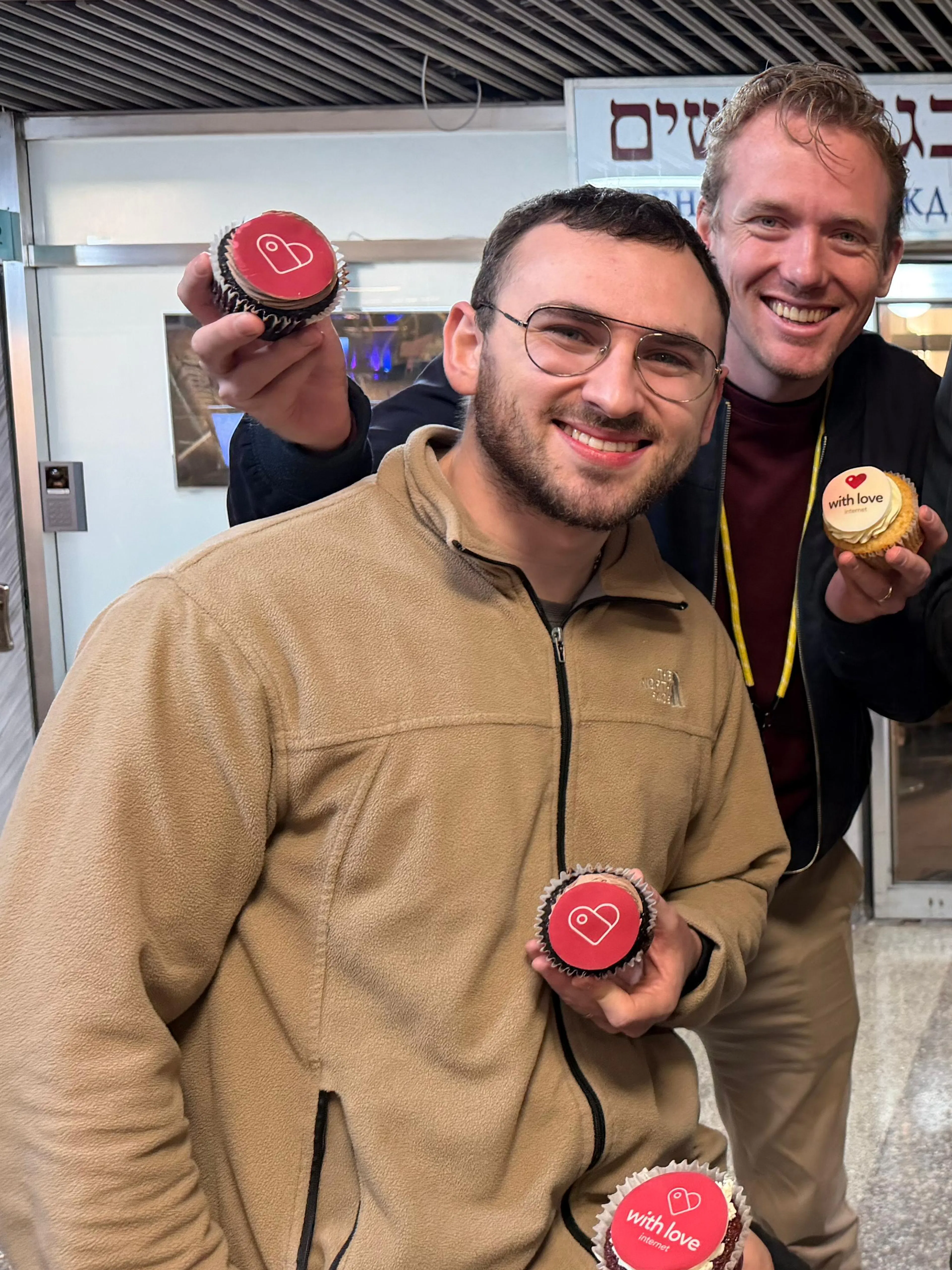 A group of five adults posing indoors, each holding a cupcake with red or yellow frosting; one man in front is about to bite into his cupcake as the others smile at the camera, with Hebrew signs visible in the background.
