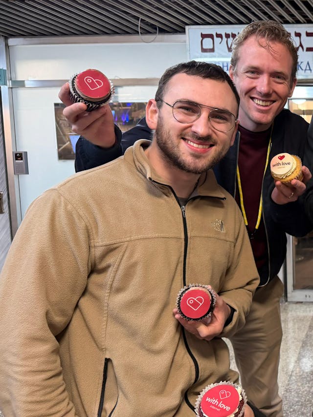 A group of five adults posing indoors, each holding a cupcake with red or yellow frosting; one man in front is about to bite into his cupcake as the others smile at the camera, with Hebrew signs visible in the background.