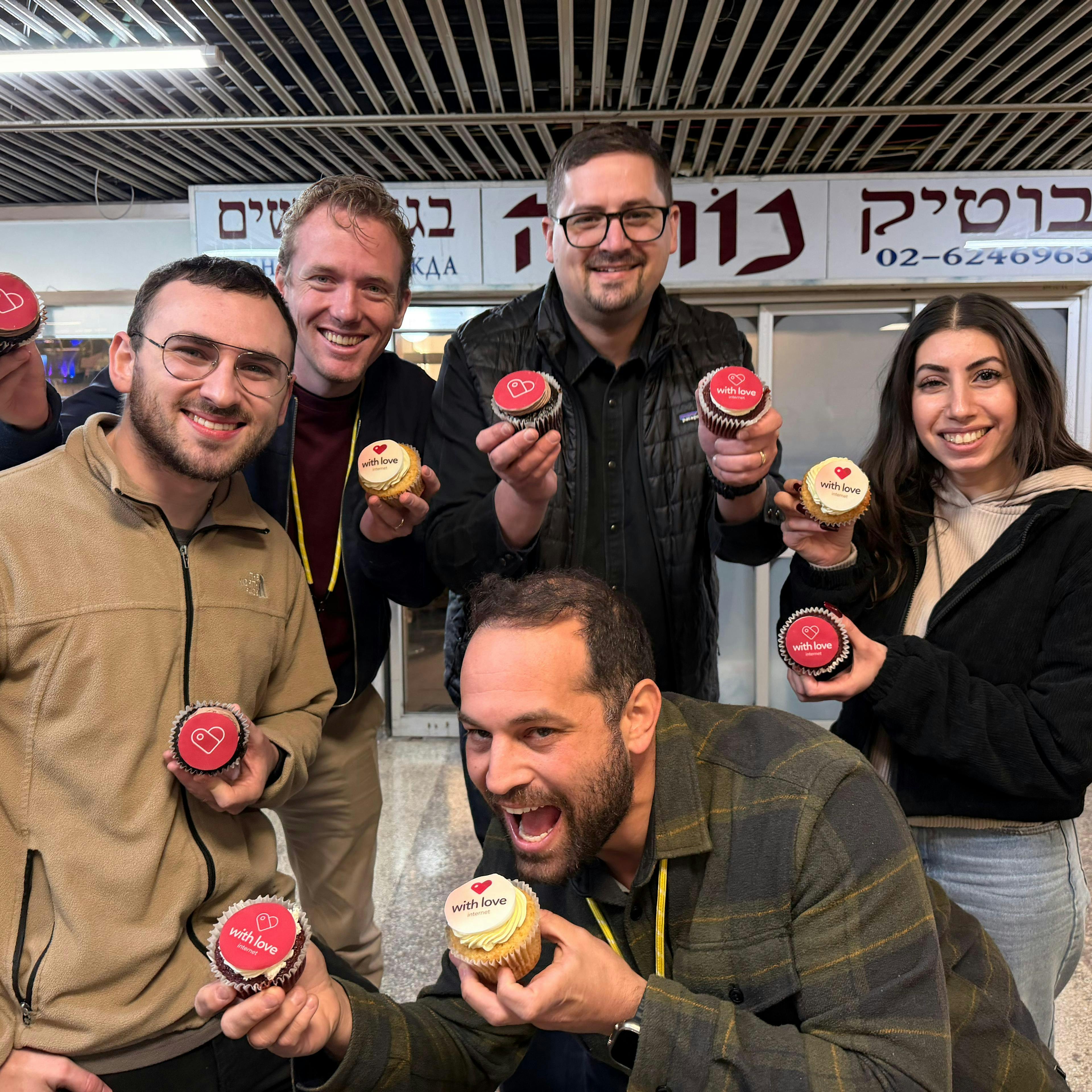 A group of five adults posing indoors, each holding a cupcake with red or yellow frosting; one man in front is about to bite into his cupcake as the others smile at the camera, with Hebrew signs visible in the background.