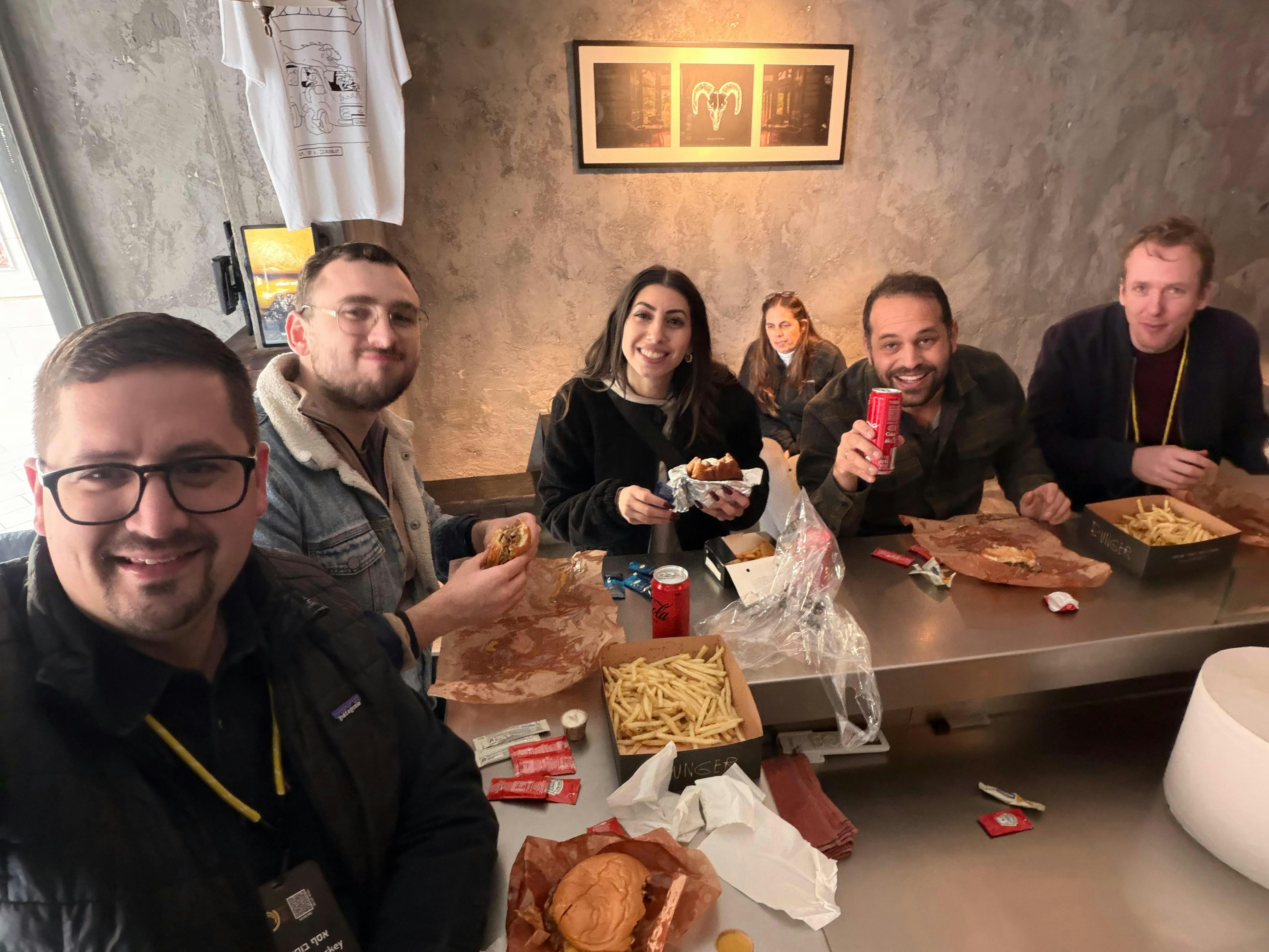 Six friends sit around a metal counter in a casual eatery, sharing burgers and fries, with drinks nearby and smiling at the camera.