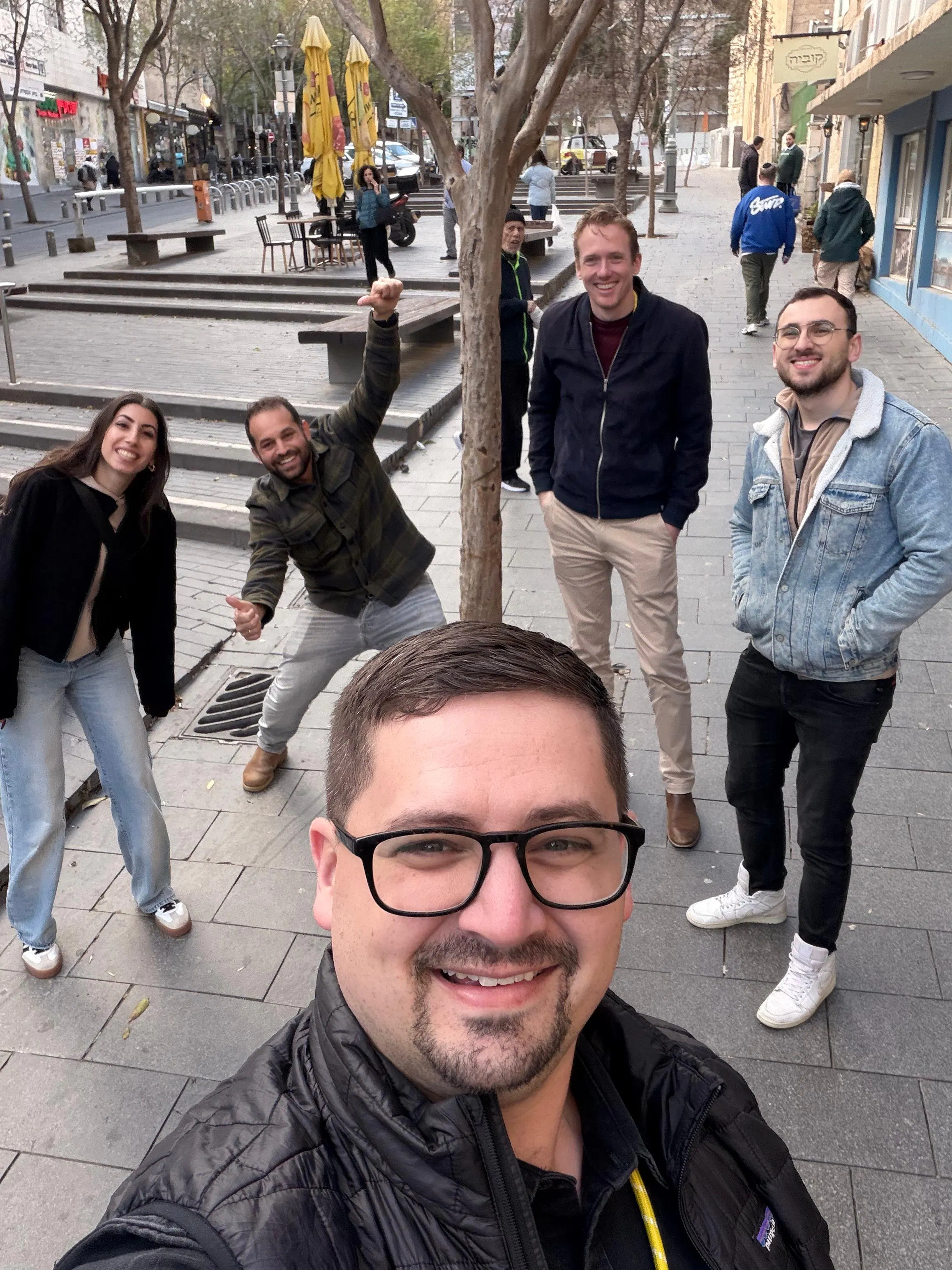 Selfie in a city plaza: a man with glasses and a black jacket smiles at the camera up front, while four friends pose behind him—a woman on the left, a man in a green jacket raising his arm, and two men in casual jackets. Trees, benches, and yellow umbrellas line the square.