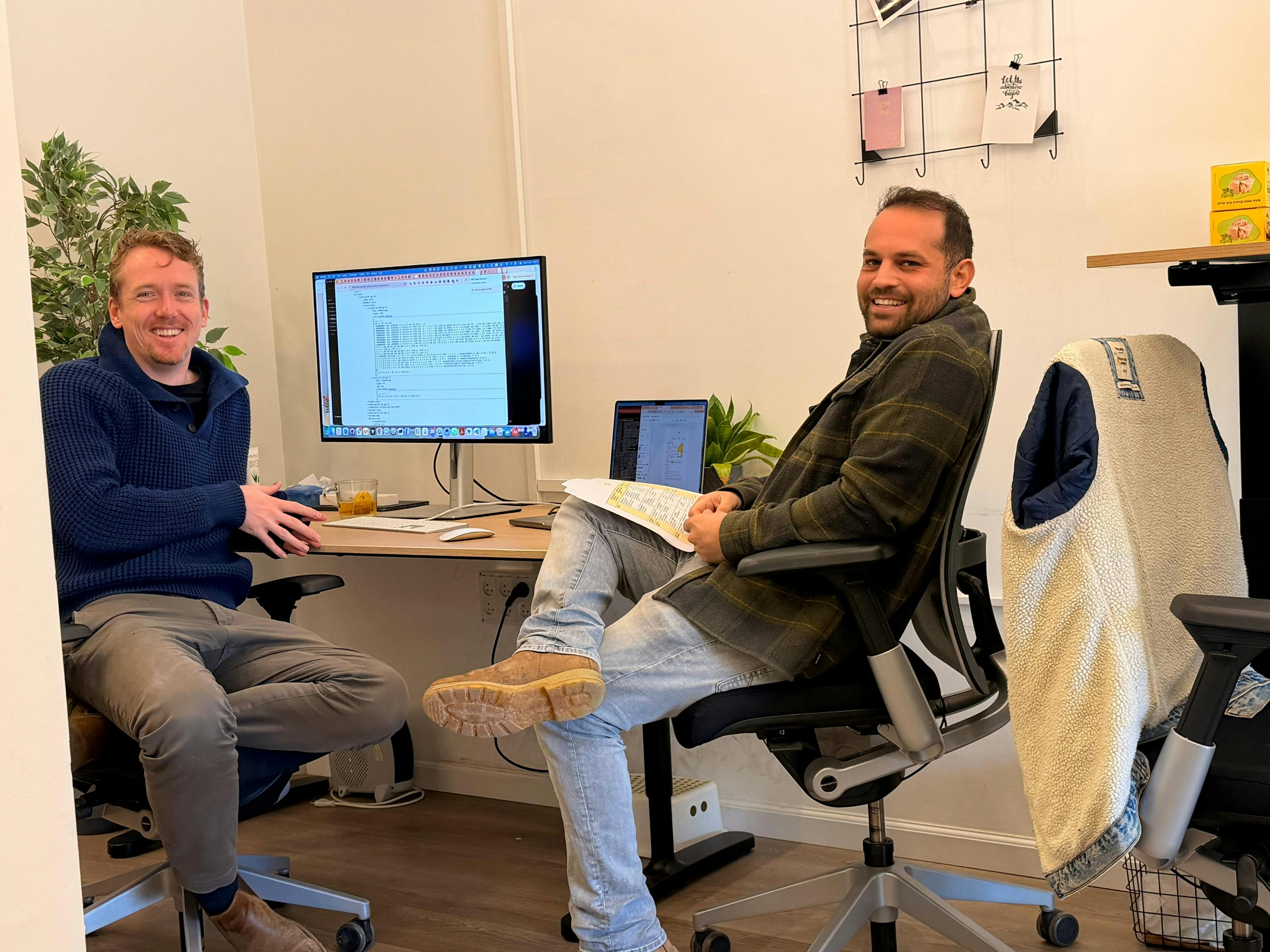 Two men sit at a small office desk smiling at the camera. One wears a blue sweater, the other a plaid shirt and jeans while holding papers. A computer monitor and laptop sit on the desk, with a plant, a jacket over a chair, and a corkboard on the wall behind.