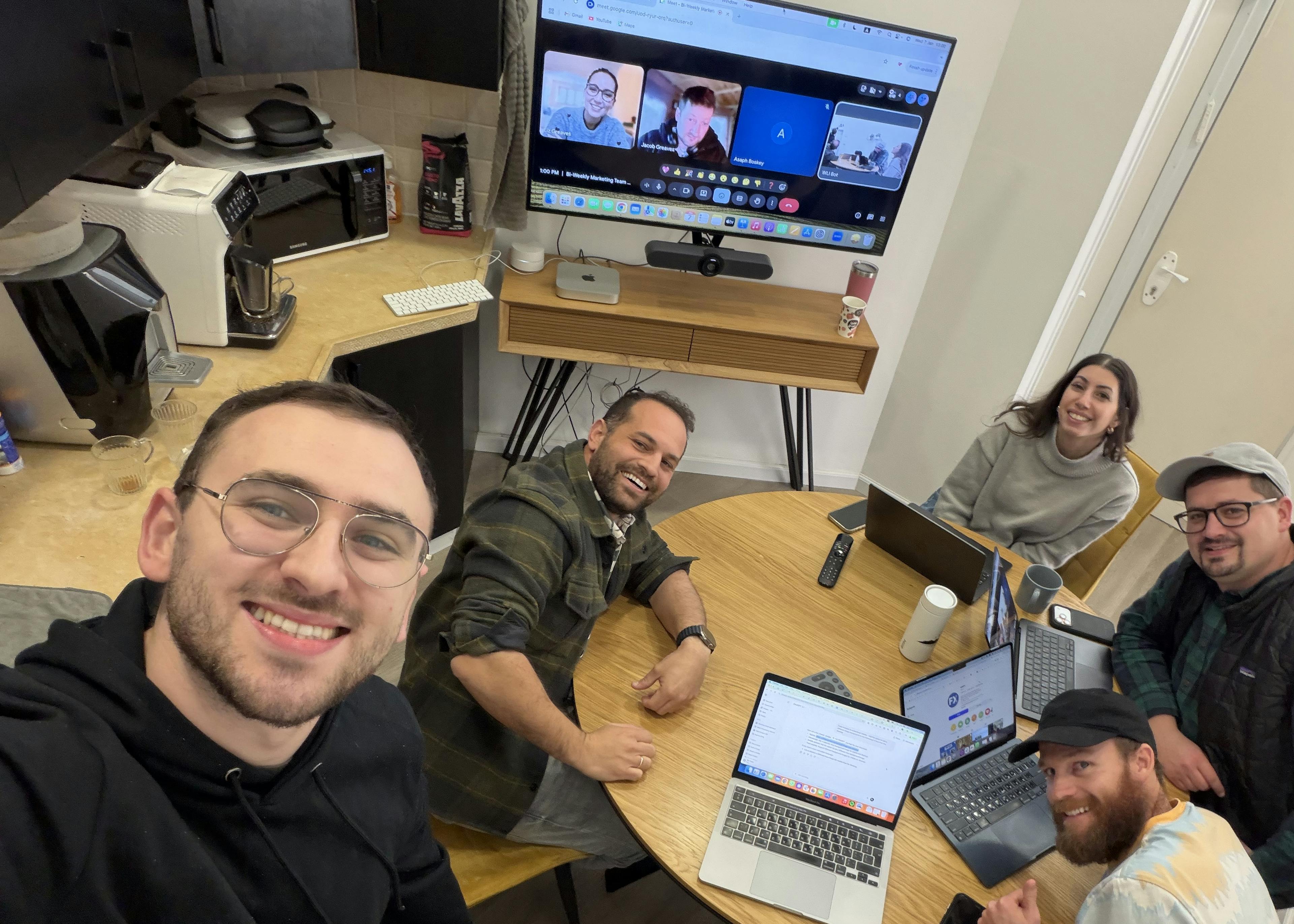 Five colleagues sit around a round wooden table in a bright office, smiling for a selfie. Laptops are open, a large monitor on a stand shows a video conference, and coffee cups sit nearby.