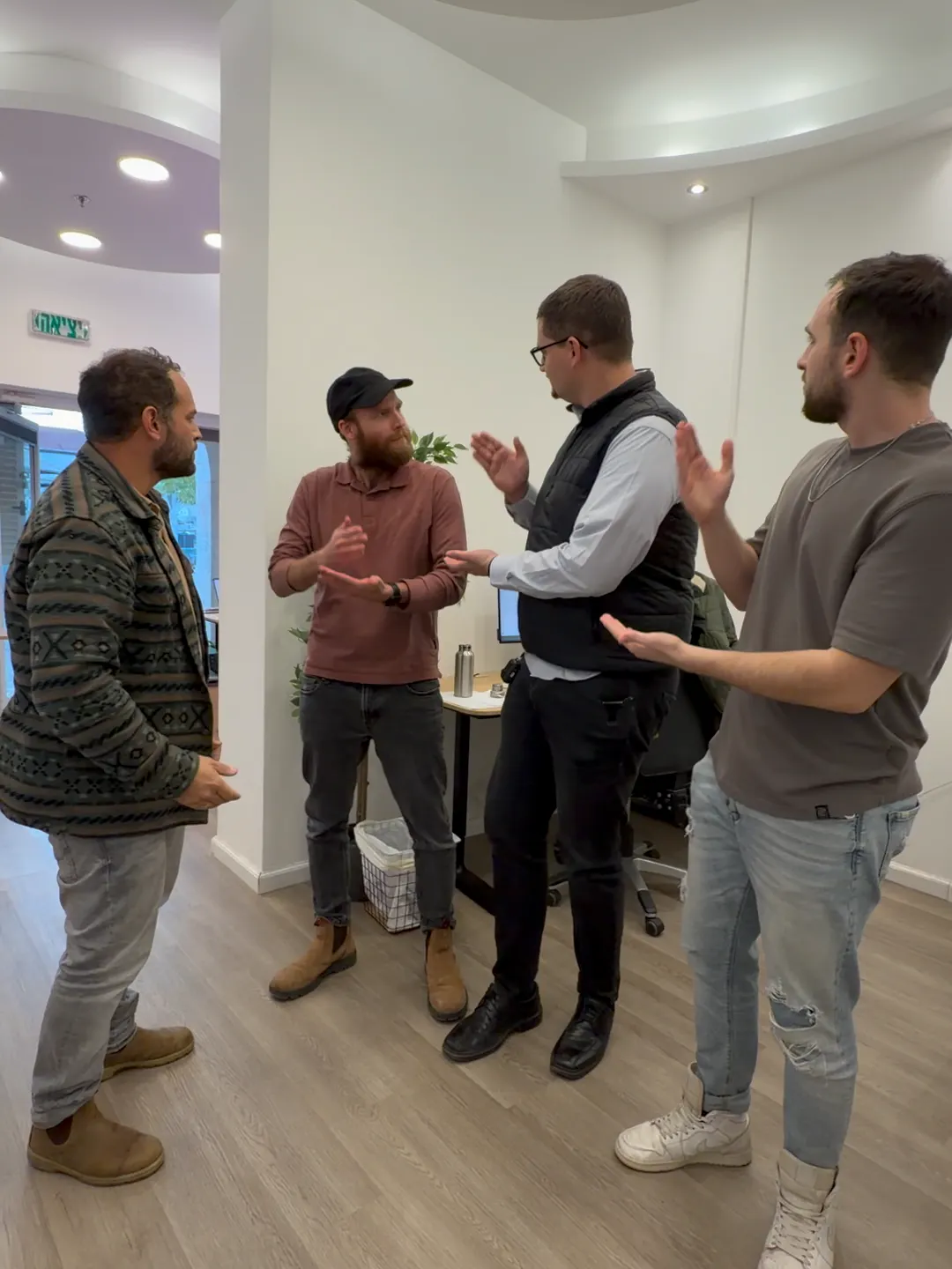Five men stand in a bright, modern room with curved white walls and circular ceiling lights. They chat in a loose circle; a man in a black vest gestures as the others listen, suggesting a casual business discussion or presentation.