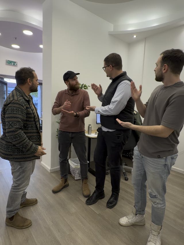 Five men stand in a bright, modern room with curved white walls and circular ceiling lights. They chat in a loose circle; a man in a black vest gestures as the others listen, suggesting a casual business discussion or presentation.