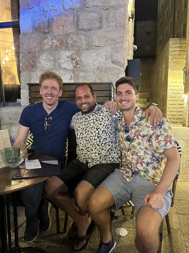 Three men sit close together at an outdoor cafe along a stone wall, smiling at the camera. They wear casual summer shirts and shorts; drinks and a bottle sit on a round table in front of them. Warm lighting and a stone staircase create a relaxed, urban-night vibe.