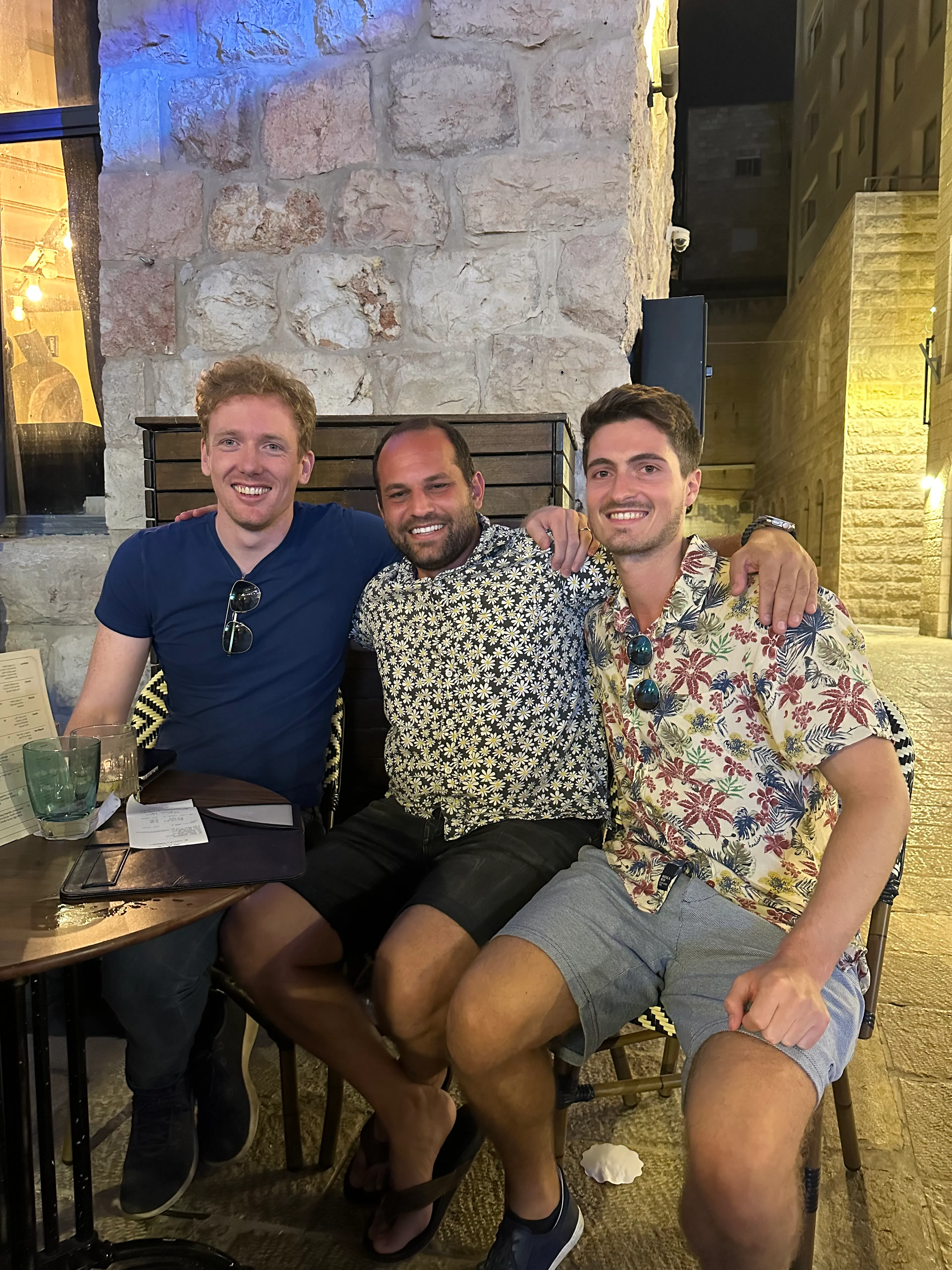 Three men sit close together at an outdoor cafe along a stone wall, smiling at the camera. They wear casual summer shirts and shorts; drinks and a bottle sit on a round table in front of them. Warm lighting and a stone staircase create a relaxed, urban-night vibe.