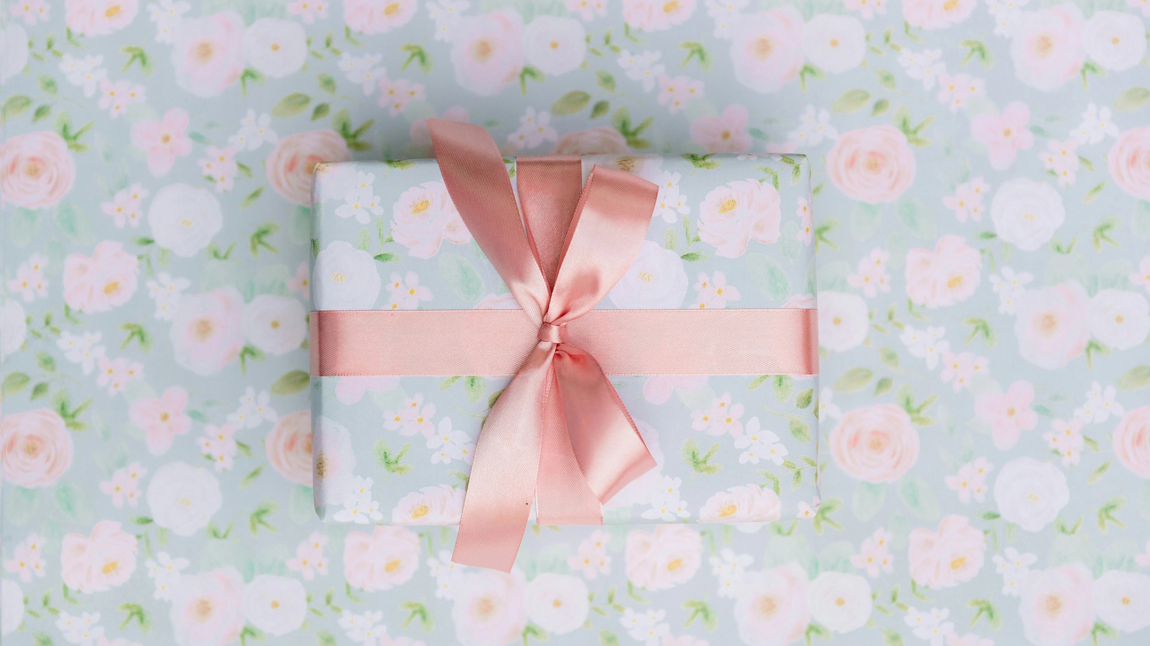 Auf einem hellblau geblümten Hintergrund liegt ein Geschenk, das mit dem selben Blümchenmuster und einer rosa Schleife verpackt ist. 