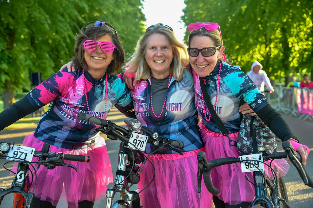 Three women with their arms round each other on their bikes, in Ride the Night gear.