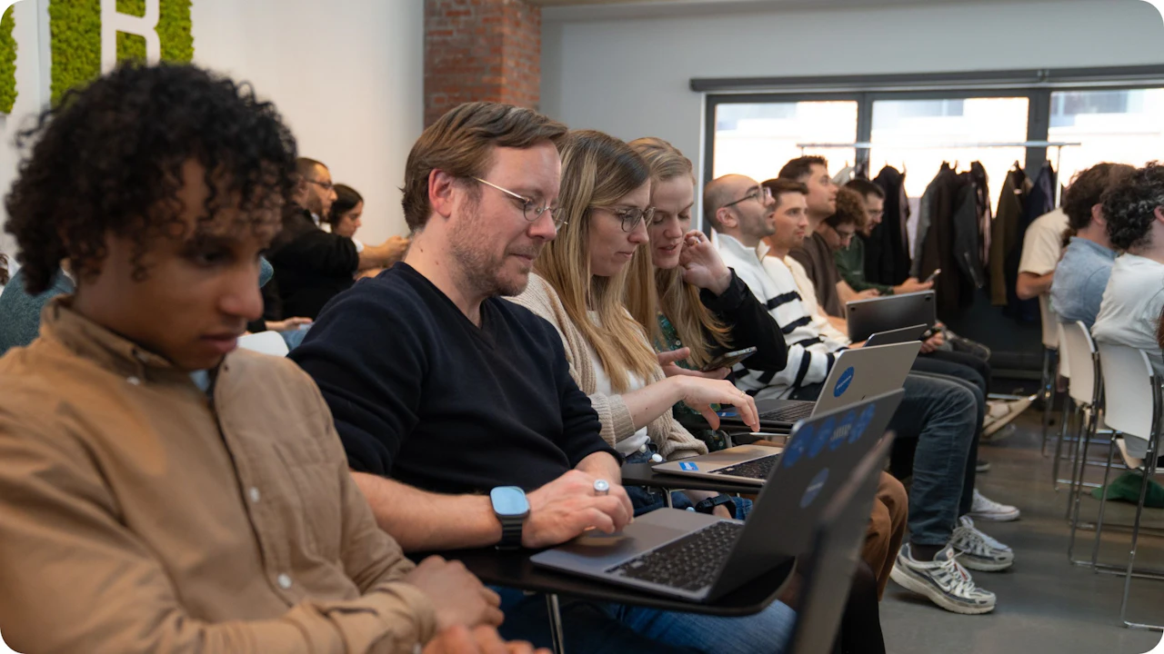 Rows of attendees seated at desks use laptops and phones during a session, with several people looking at the same screen together.