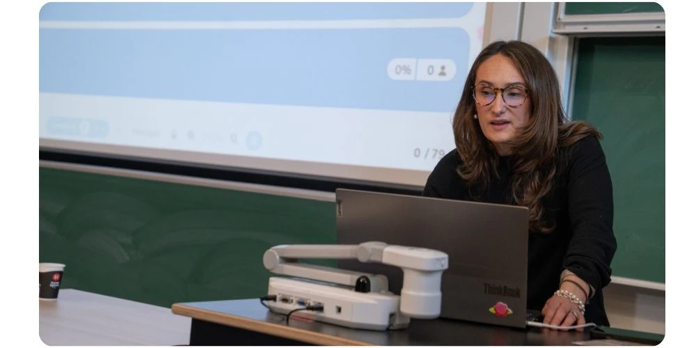 A teacher sits at a desk with a laptop and a small robotic arm, speaking to a class while a Wooclap results screen showing 0% and 0 participants is projected behind her