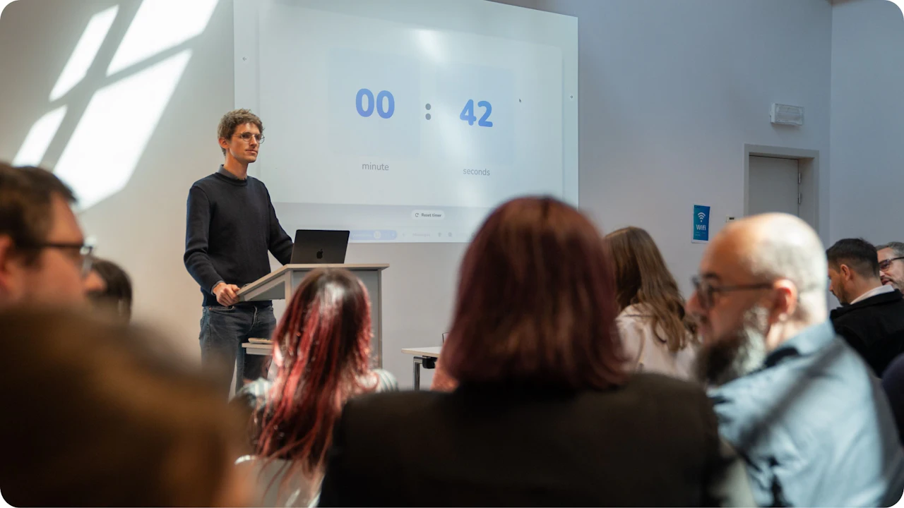 A presenter stands at a podium with a laptop beside a projected Wooclap countdown timer showing 00:42, while attendees watch from the front rows.