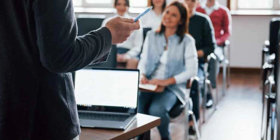 Guru mengajar dengan laptop di depannya, sementara para siswa mendengarkan.