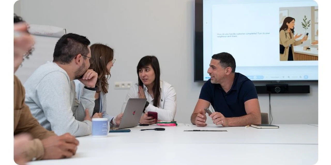 A group of colleagues sit around a table talking, with a screen behind them displaying a Wooclap discussion prompt about handling customer complaints