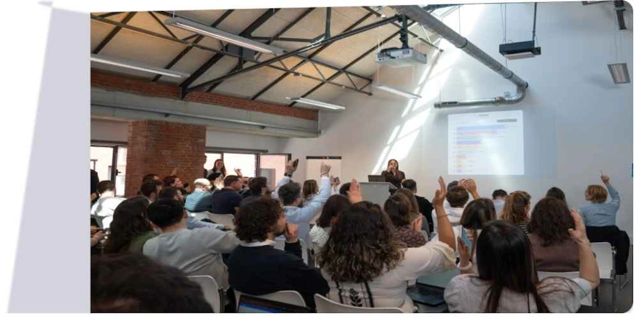 A large group of participants raising their hands during an interactive presentation, with a Wooclap activity displayed on the screen at the front of the room.