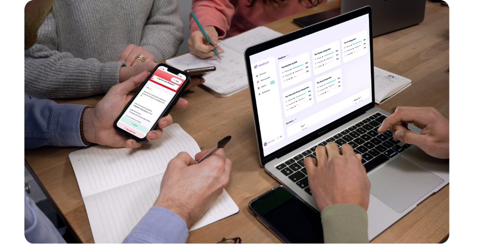 People taking notes at a table while checking training progress on a laptop showing the Wooflash dashboard and a phone displaying a Wooflash flashcard.