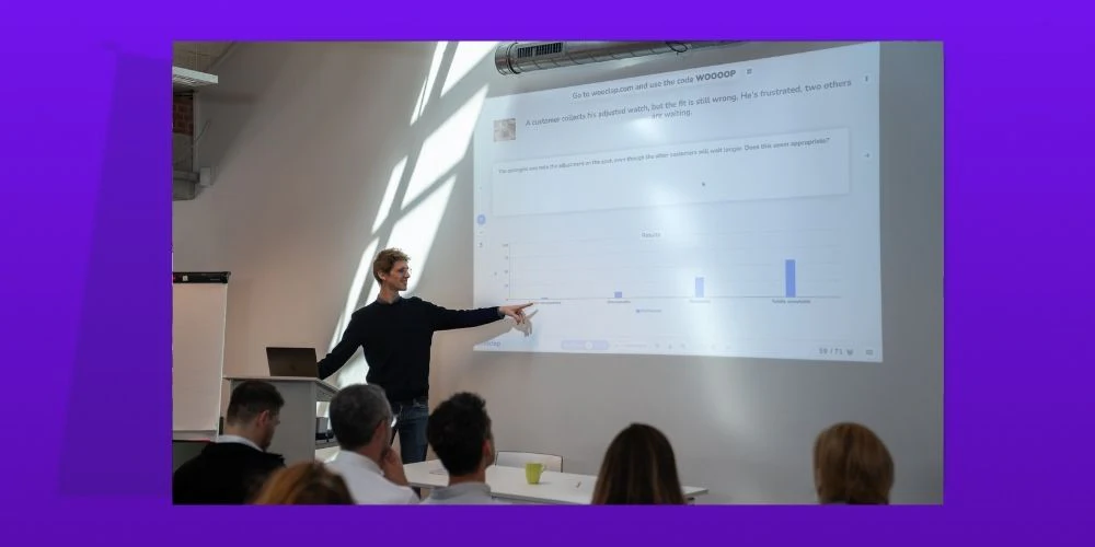 Ein Präsentator steht an einem Podium und zeigt auf eine Projektionsleinwand, die eine Wooclap-Umfrage mit einer Frage zu einem Kundenservice-Szenario und ein Balkendiagramm der Publikumsantworten zeigt, während mehrere Teilnehmer an Tischen zuschauen.