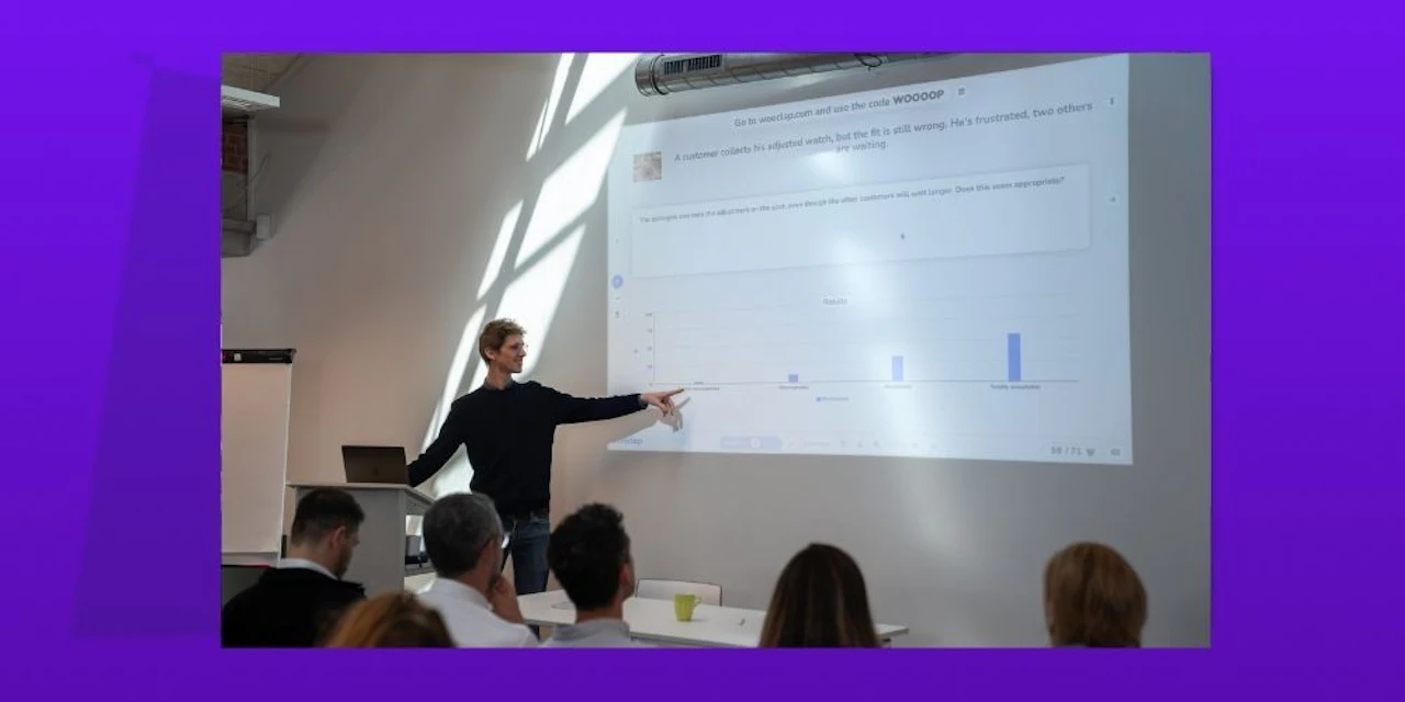 Ein Präsentator steht an einem Podium und zeigt auf eine Projektionsleinwand, die eine Wooclap-Umfrage mit einer Frage zu einem Kundenservice-Szenario und ein Balkendiagramm der Publikumsantworten zeigt, während mehrere Teilnehmer an Tischen zuschauen.