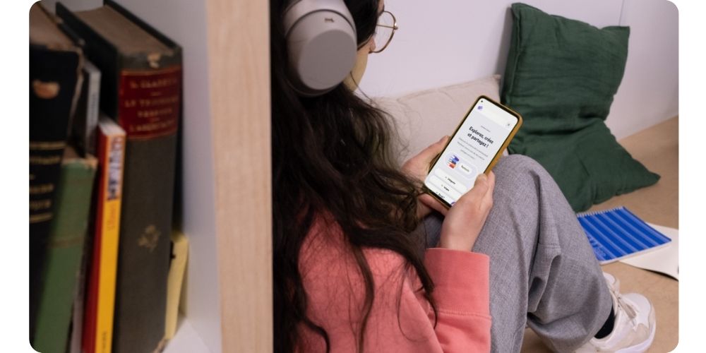 A woman wearing headphones sits on a couch browsing the Wooclap mobile app on her phone, with bookshelves in the background.
