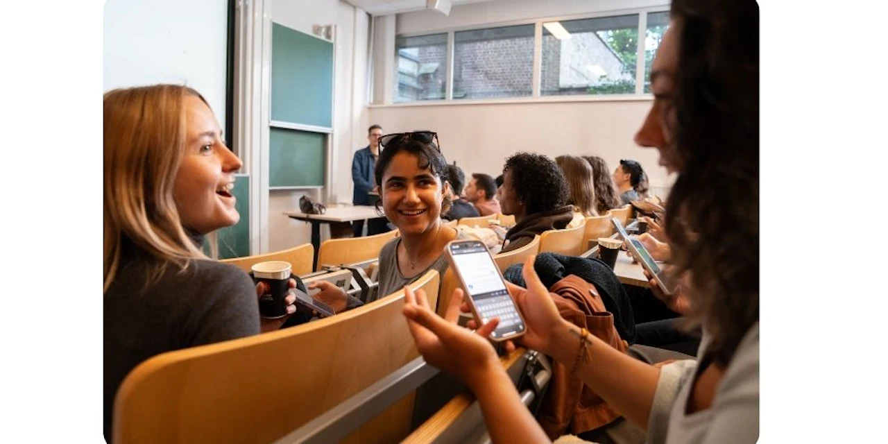 Three students smile and talk together in a lecture hall while holding coffee cups and smartphones with a Wooclap session open.