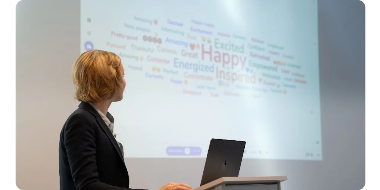 Una relatrice al leggio, con un laptop, guarda uno schermo di proiezione che mostra una nuvola di parole di Wooclap con parole come Happy, Inspired, Excited ed Energized.