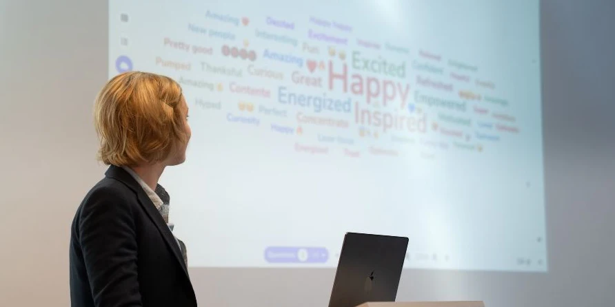 Una relatrice al leggio, con un laptop, guarda uno schermo di proiezione che mostra una nuvola di parole di Wooclap con parole come Happy, Inspired, Excited ed Energized.