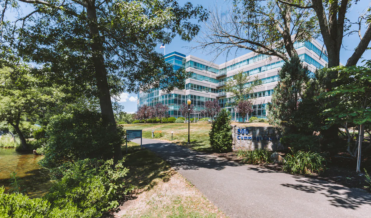 Trees and a walking path in the foreground with a sign for 500 Cummings Center. The building is in the background.