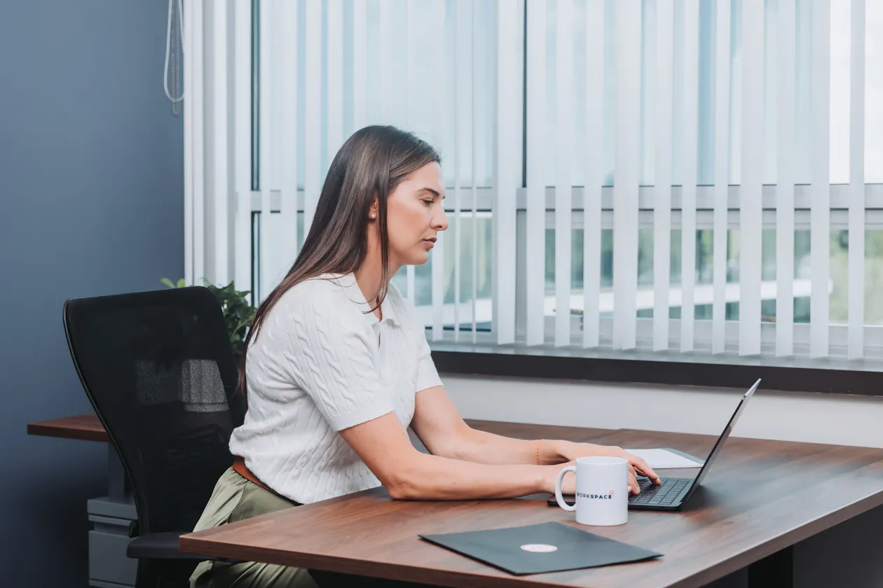 Woman sitting at a desk in an office at Workspace. She is working on a laptop and there is a coffee cup and folder on the desk.