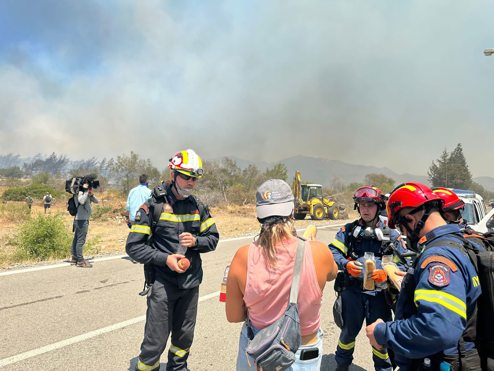 A woman in a pink shirt hands out fruit to firefighters with smoke visible in the background.