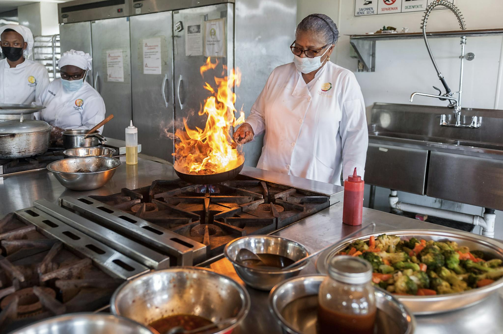 World Central Kitchen in Action: A person is standing in a industry-grade kitchen in front of a burning frying pan. Two other persons are watching astonishedly.