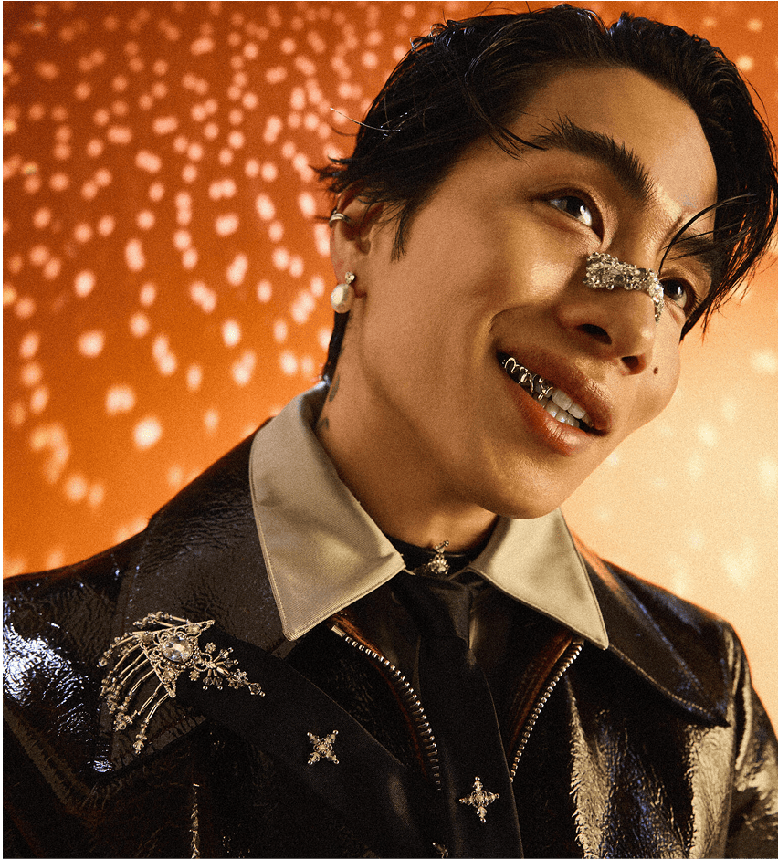 Artist SIRUP smiling while wearing a stylish outfit, adorned with accessories, against a warm orange backdrop.