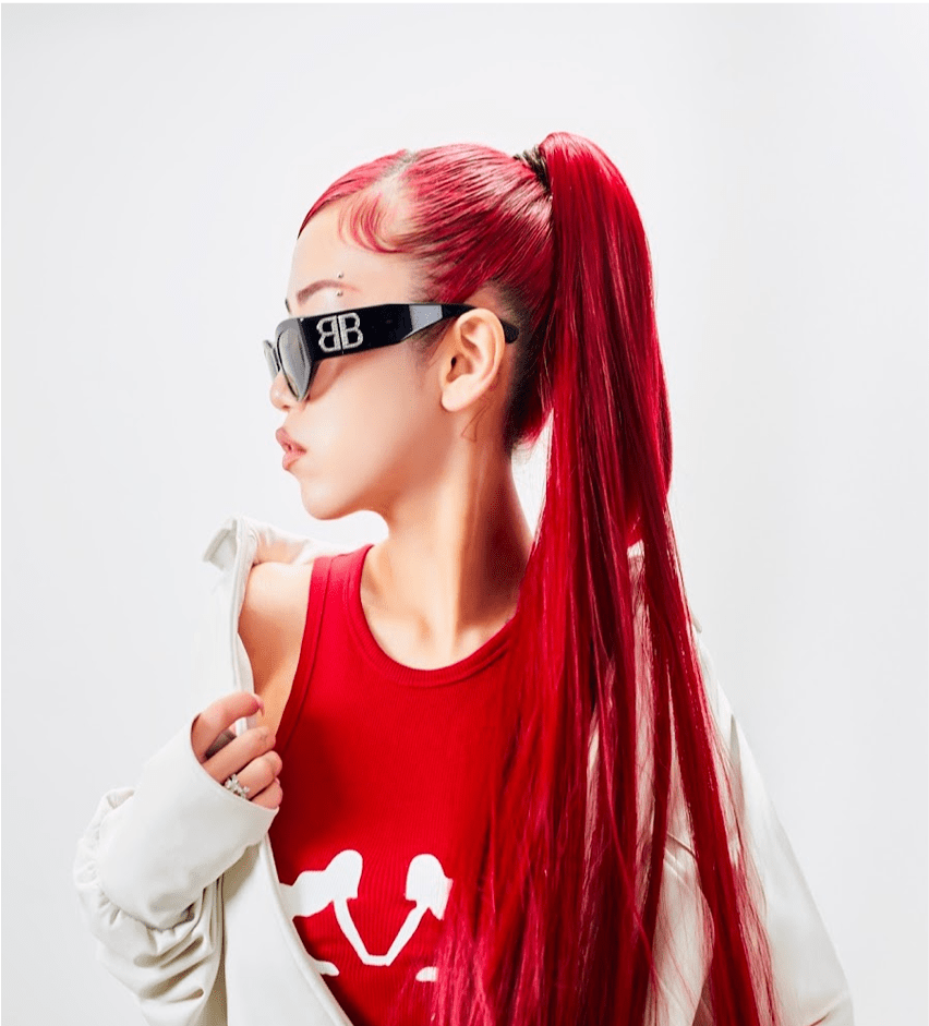 A person with long red hair styled in a ponytail and wearing dark sunglasses poses confidently in a red tank top and white jacket against a plain background. The scene captures a vibrant and stylish moment, aligning with a music event theme.