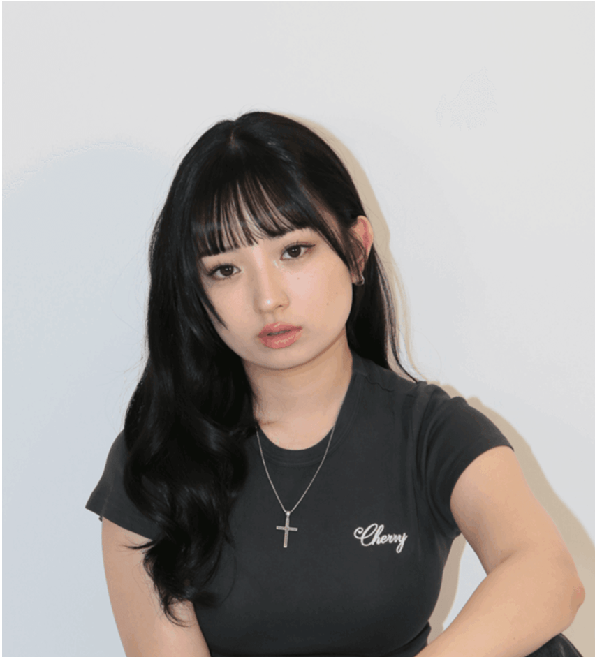 A young woman with long black hair and bangs sits thoughtfully, wearing a dark shirt with 'Cherry' printed on it, against a plain white background.