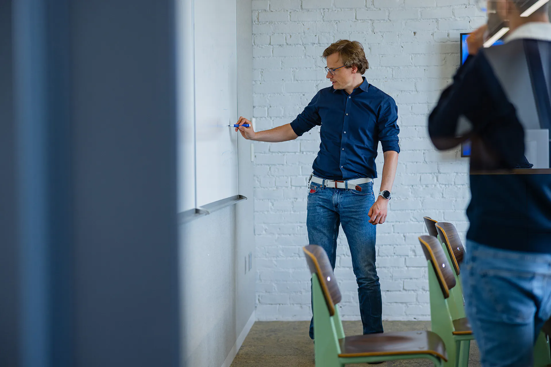 A person standing at a whiteboard, writing notes during a presentation in a bright room with chairs in the foreground.