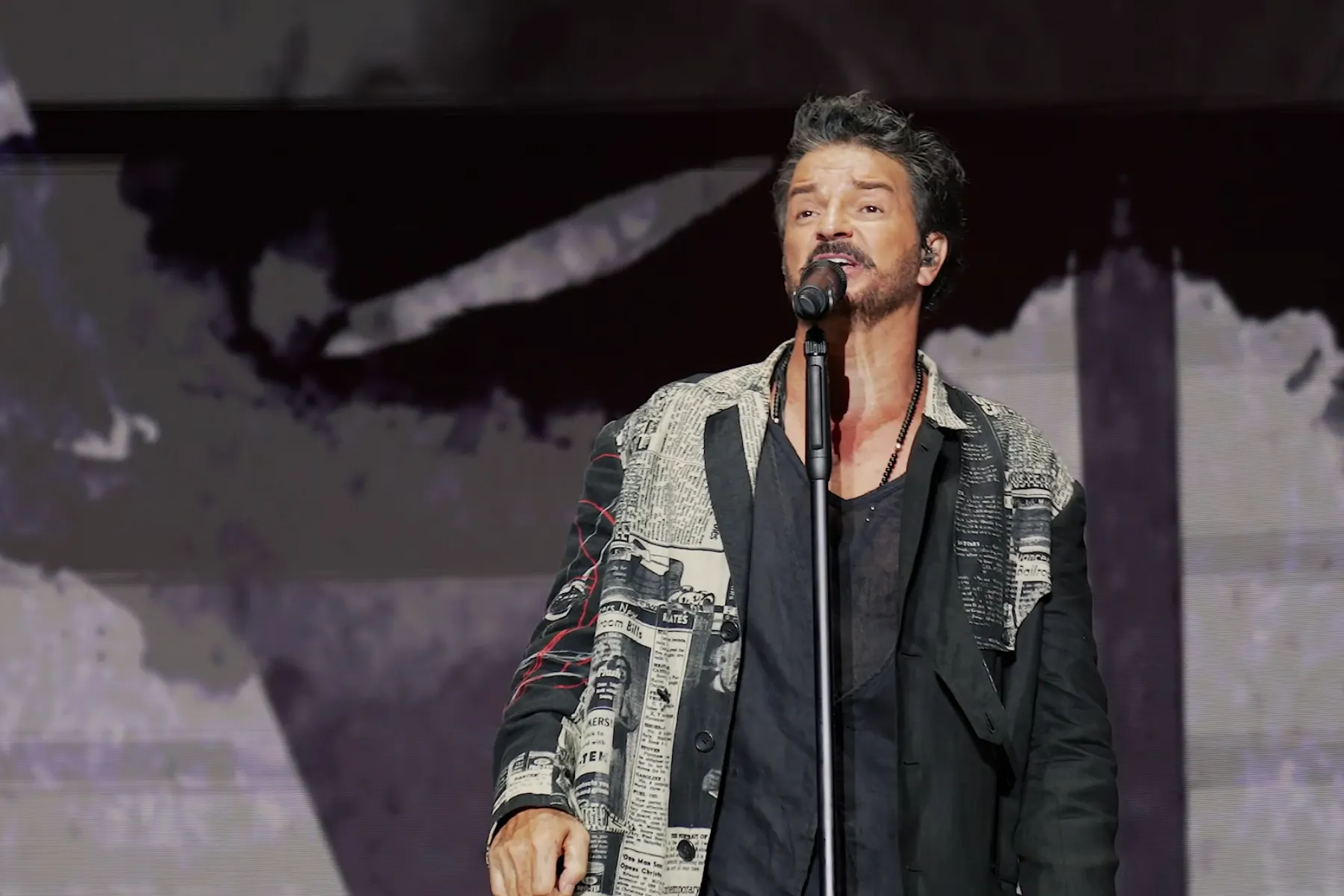 A performer singing passionately into a microphone on stage, wearing a stylish jacket with a textured design. The background features abstract visuals, enhancing the energetic atmosphere.