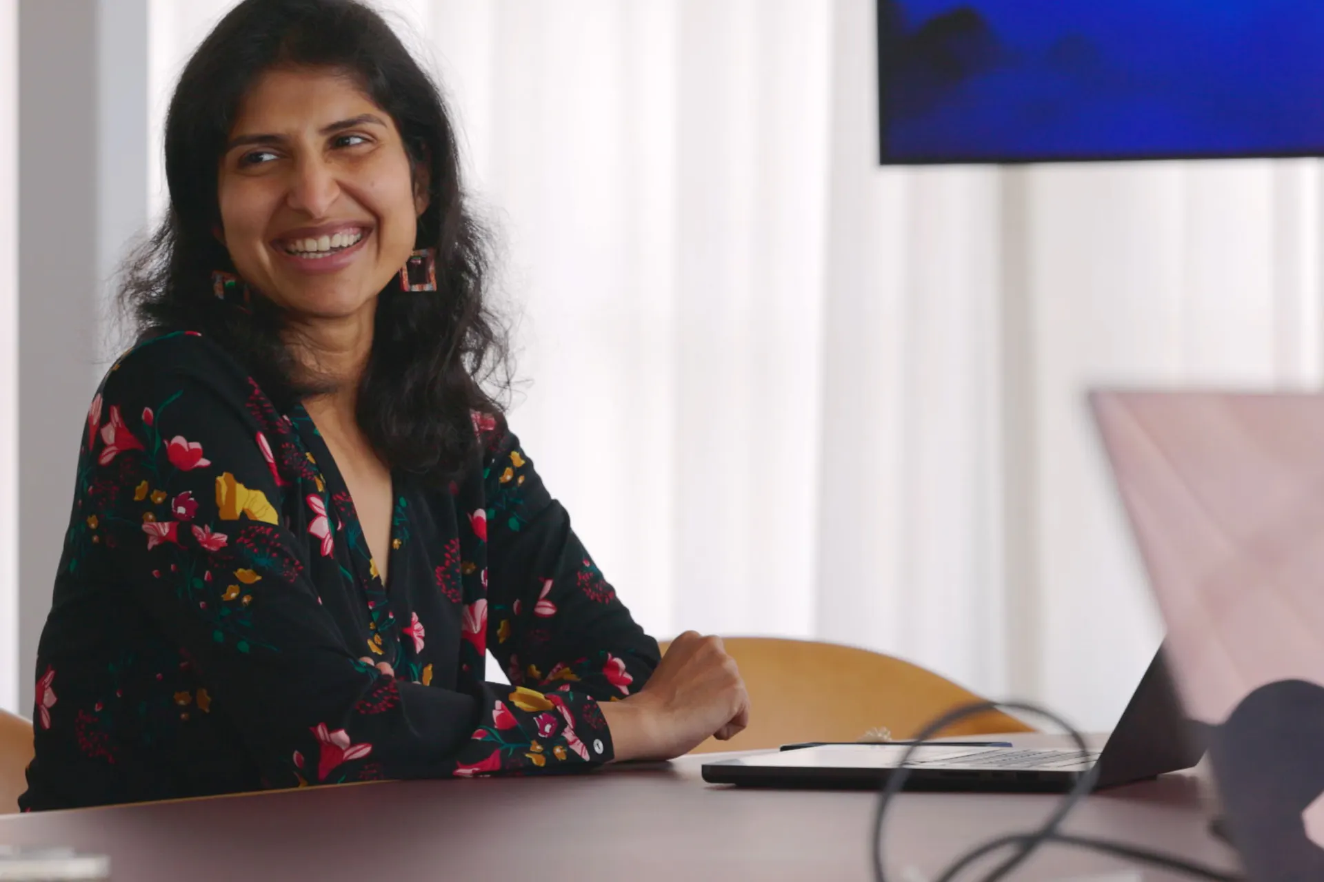 A woman with long black hair and a floral dress smiles while seated at a table with a laptop, suggestive of an engaging business discussion.