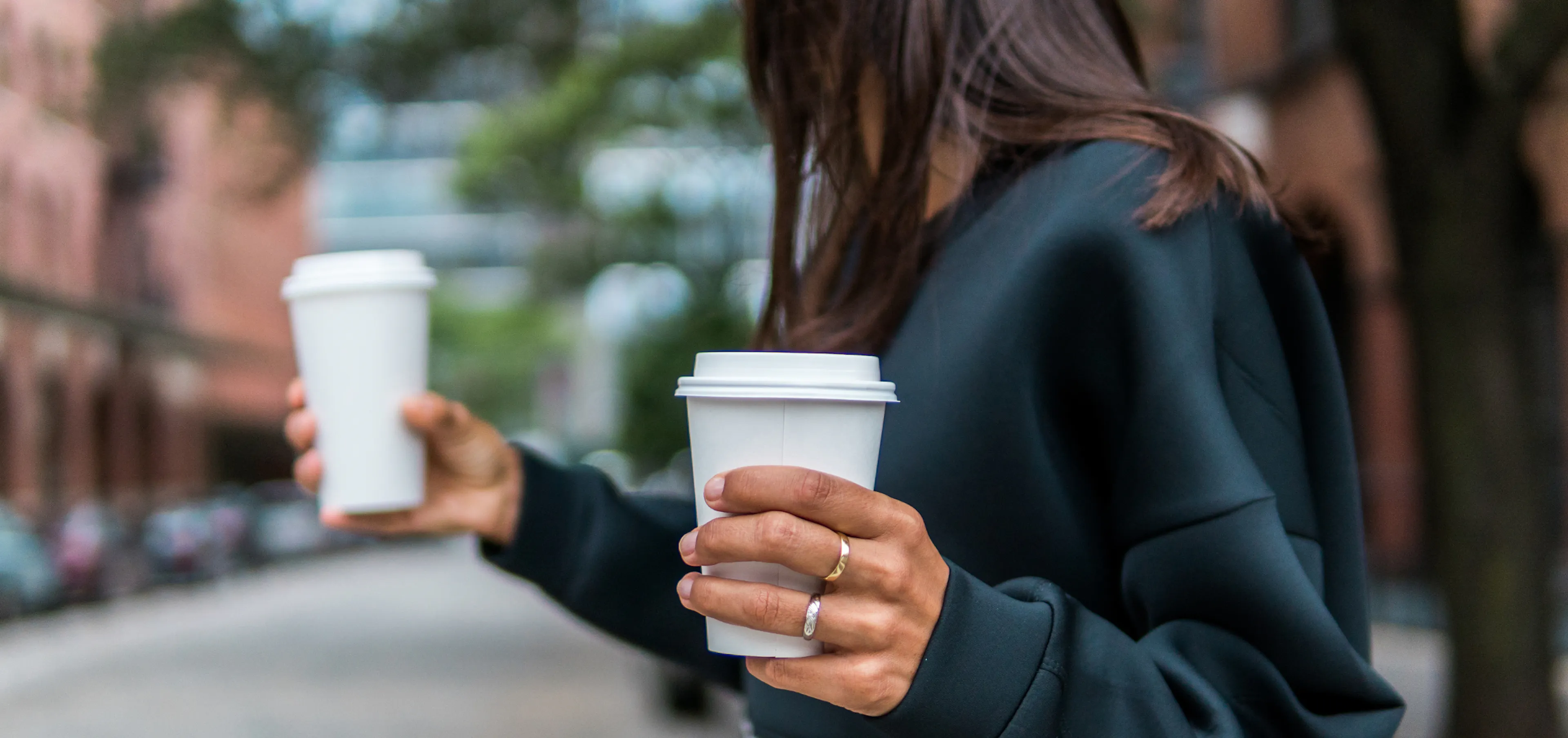 Woman holding two takeout coffees on a city sidewalk, reaching out to hand one to someone as a small gesture of appreciation